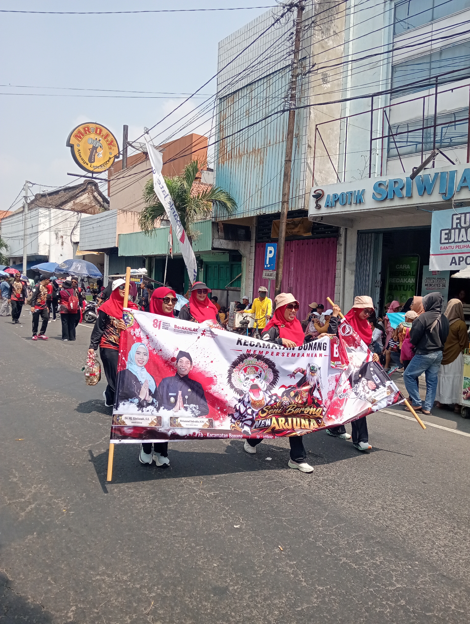 The image captures a daytime street scene in Demak, Indonesia, likely during a parade or procession. In the foreground, a group of people, primarily women wearing red headscarves and some in matching black and red outfits, are holding a large banner. The banner features colorful graphics, including images of two individuals, stylized masks, and text in Indonesian. One of the individuals on the banner is identified as "Dr. Hj. Elstianah, S.E." and the other as "Muhammad Rofikhudin, M.M.". The banner also has text that appears to read "Kecamatan Bonang" and "Seni Barong NEW ARJUNA". To the left of the banner, one woman holds a basket containing small items. In the background, other participants in the procession are visible, some carrying flags. Buildings line the street, including one with a sign that says "APOTIK SRIWIJAY" and another with a circular sign featuring a magnifying glass and the text "MR. DIY". Power lines crisscross the sky above. The street surface is asphalt. The overall atmosphere appears festive, with a clear sky overhead.