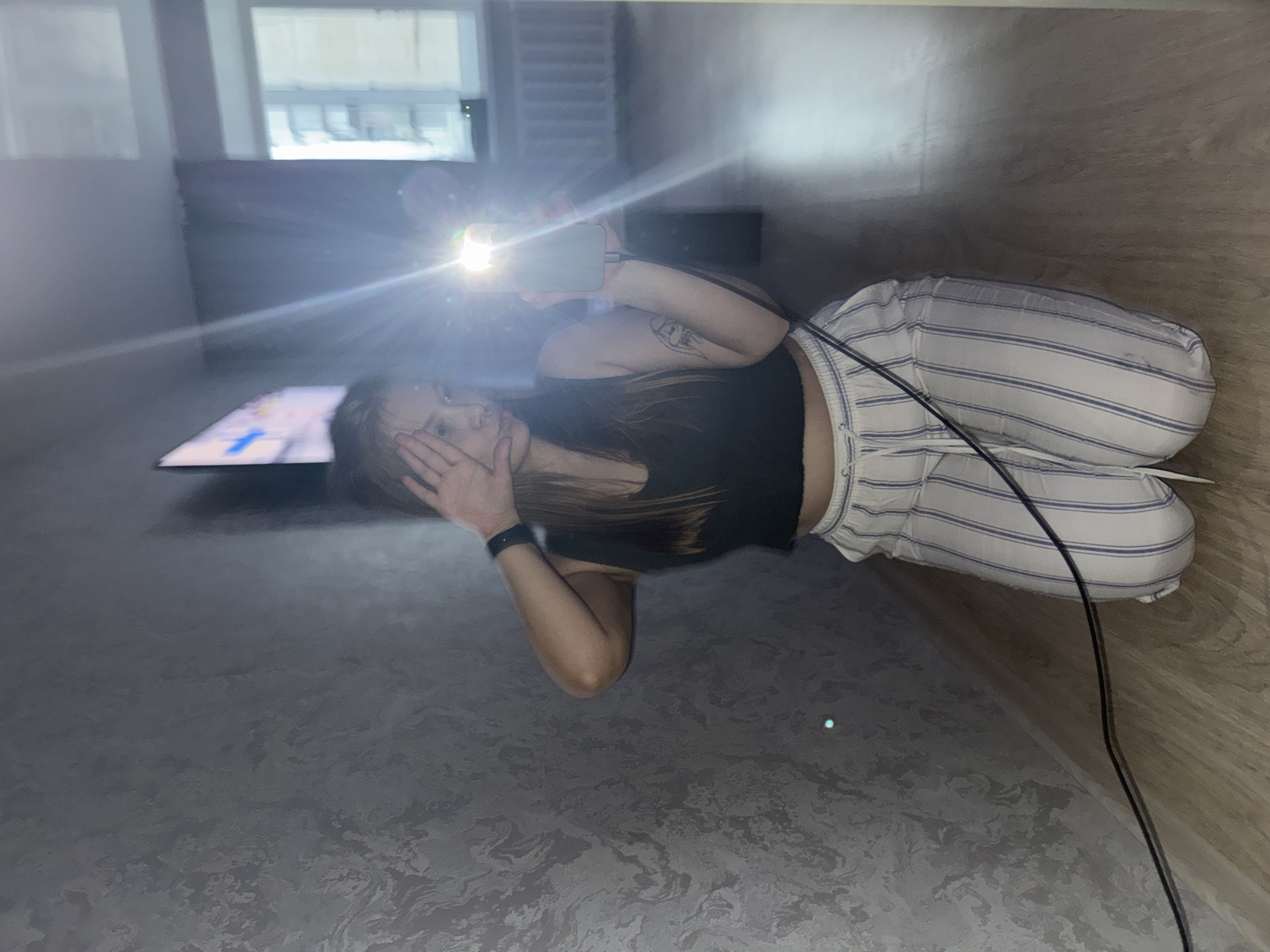 A young woman is holding up a smartphone with the flash on, covering part of her face with her hand. She is wearing a black tank top and striped pants. An electrical cord runs from the bottom right of the image. In the background is a TV screen, a window, and a sofa. The location is within a building, possibly a home, in the city of Chaikovsky, Russia. The floor has a mottled gray pattern, and the wall is paneled in wood.