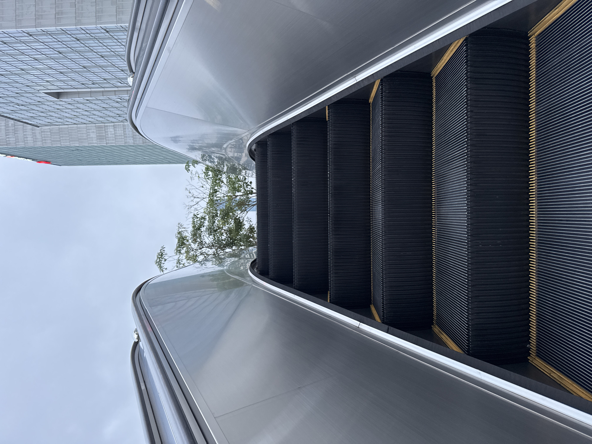 The image depicts an outdoor escalator in Futian District, China, likely connecting different levels or areas of an outdoor complex. The escalator's steps are black with yellow safety markings. The sides of the escalator are silver. A building with a grid-like facade is visible in the background on the left. There are some trees behind the escalator. The sky is overcast.