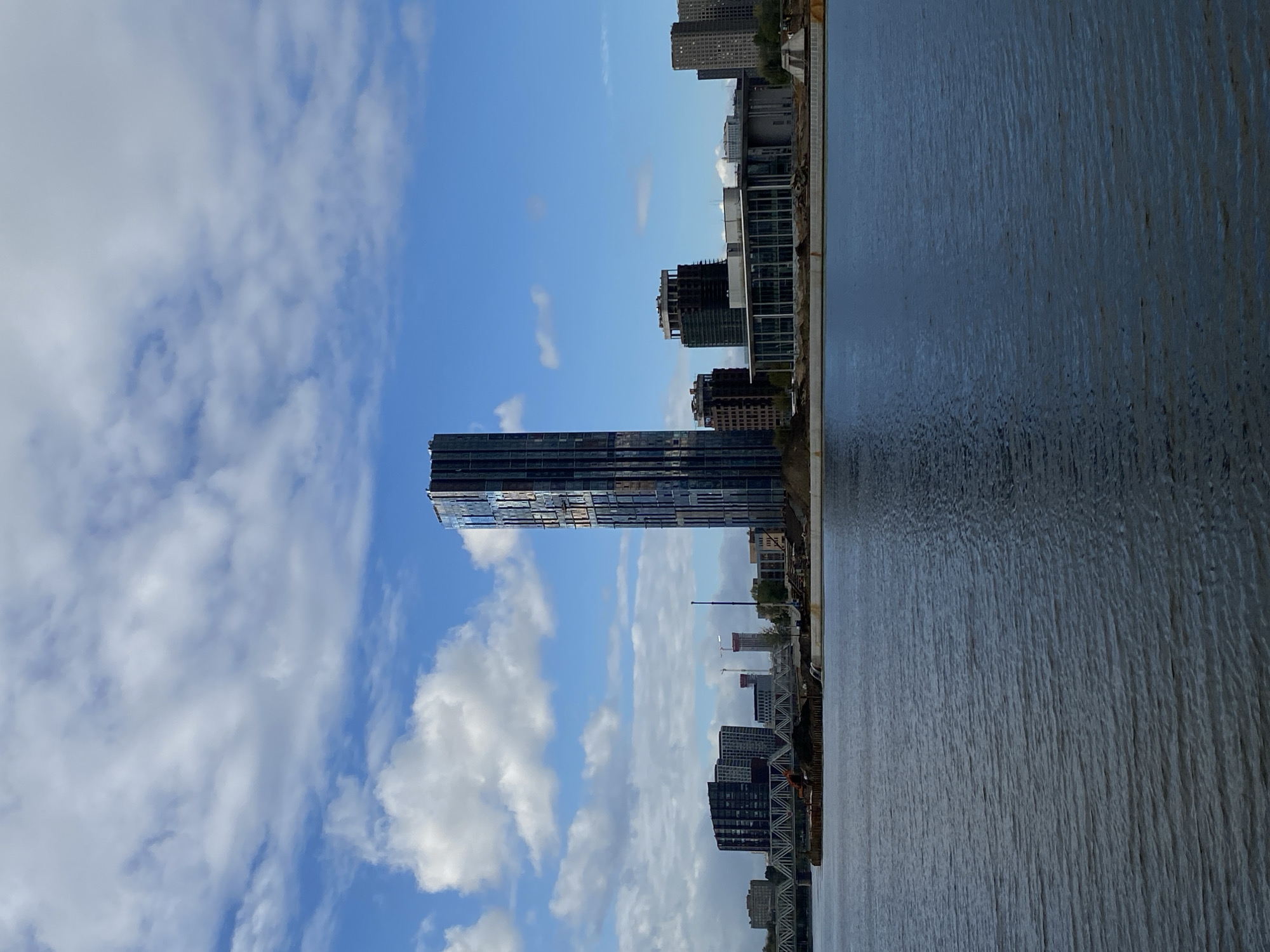 The image shows a waterfront cityscape with a prominent dark blue skyscraper with a glass facade dominating the center. Several other buildings of varying heights and architectural styles are visible in the background, including mid-rise apartment buildings and what appears to be a modern office or public building with large glass windows and a white roof. A bridge with a steel truss structure spans across the water in the distance. The foreground features a body of dark blue water with ripples, suggesting a slight breeze. Along the shoreline, there are signs of construction, with piles of dirt, debris, and some industrial equipment, including an orange excavator. The sky is a bright blue with scattered white clouds. The scene depicts a developing urban waterfront area under a partly cloudy sky.