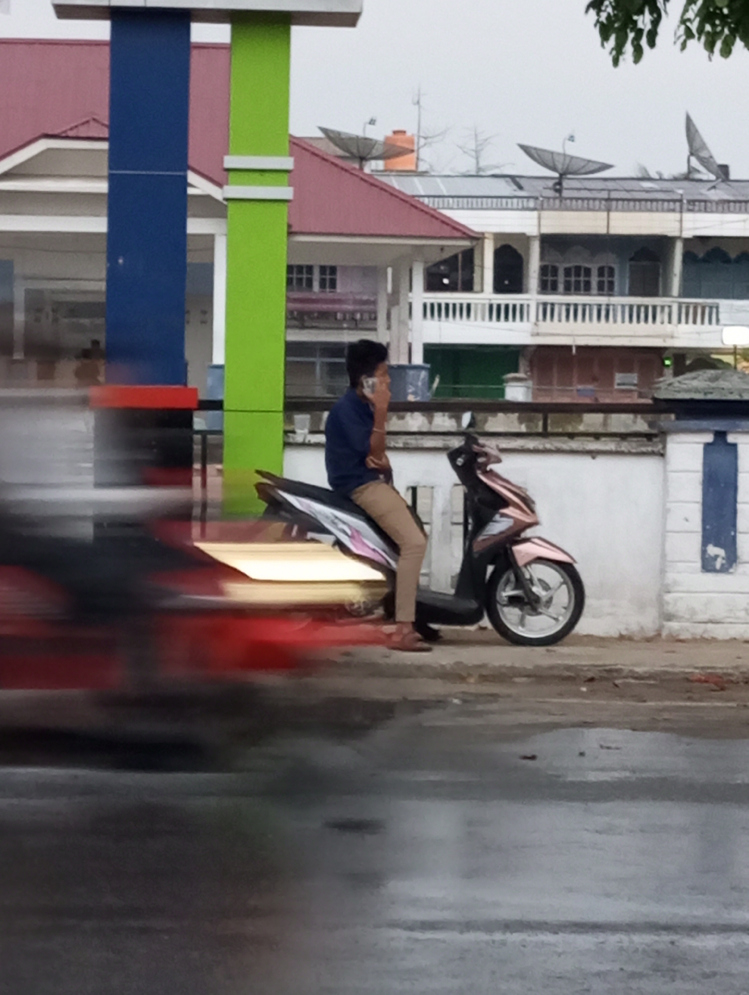 In Air Molek, Indonesia, a man is seated on a pink and black scooter. He is wearing a dark blue shirt and light brown pants, and he is holding a phone to his ear. The scooter is parked next to a white wall with blue accents. In the background, there are buildings with red roofs and satellite dishes. The ground appears wet, suggesting it may have recently rained. Blurred red and yellow streaks indicate traffic movement in the foreground.