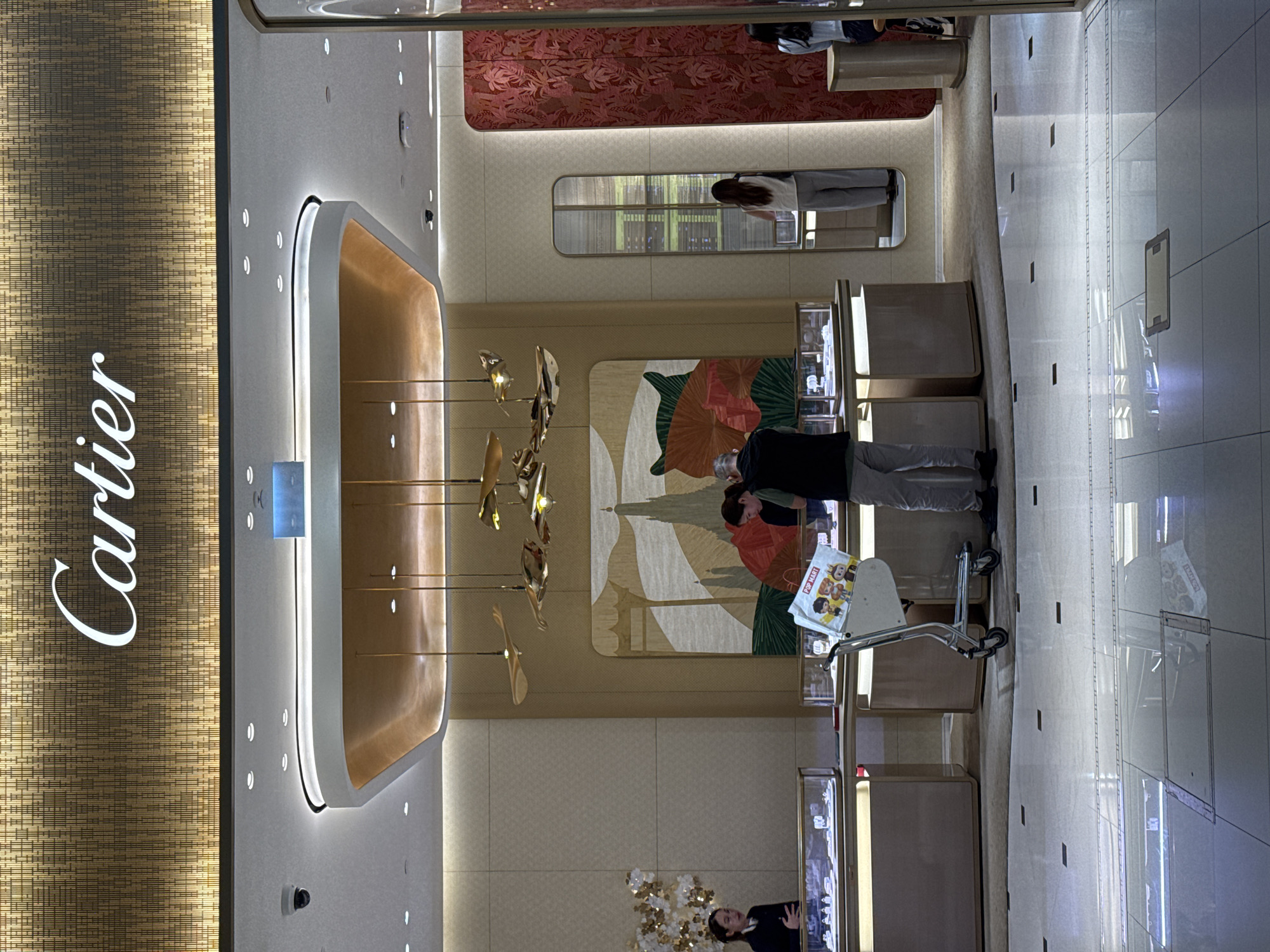 In a brightly lit, modern interior, possibly a luxury boutique, a person is seen pushing a metal shopping cart. The cart has a white bag with colorful cartoon characters and text that appears to say "POP MART". The person pushing the cart is wearing dark clothing. In the background, the store displays are visible, with illuminated shelves and glass cases. The store's signage is prominently displayed on the left side of the image, featuring the word "Cartier" in white, stylized lettering against a textured, gold-toned background. The overall ambiance suggests a retail environment, likely during daylight hours given the interior lighting. The scene appears calm and orderly. The location is identified as Nong Prue Subdistrict, Thailand.