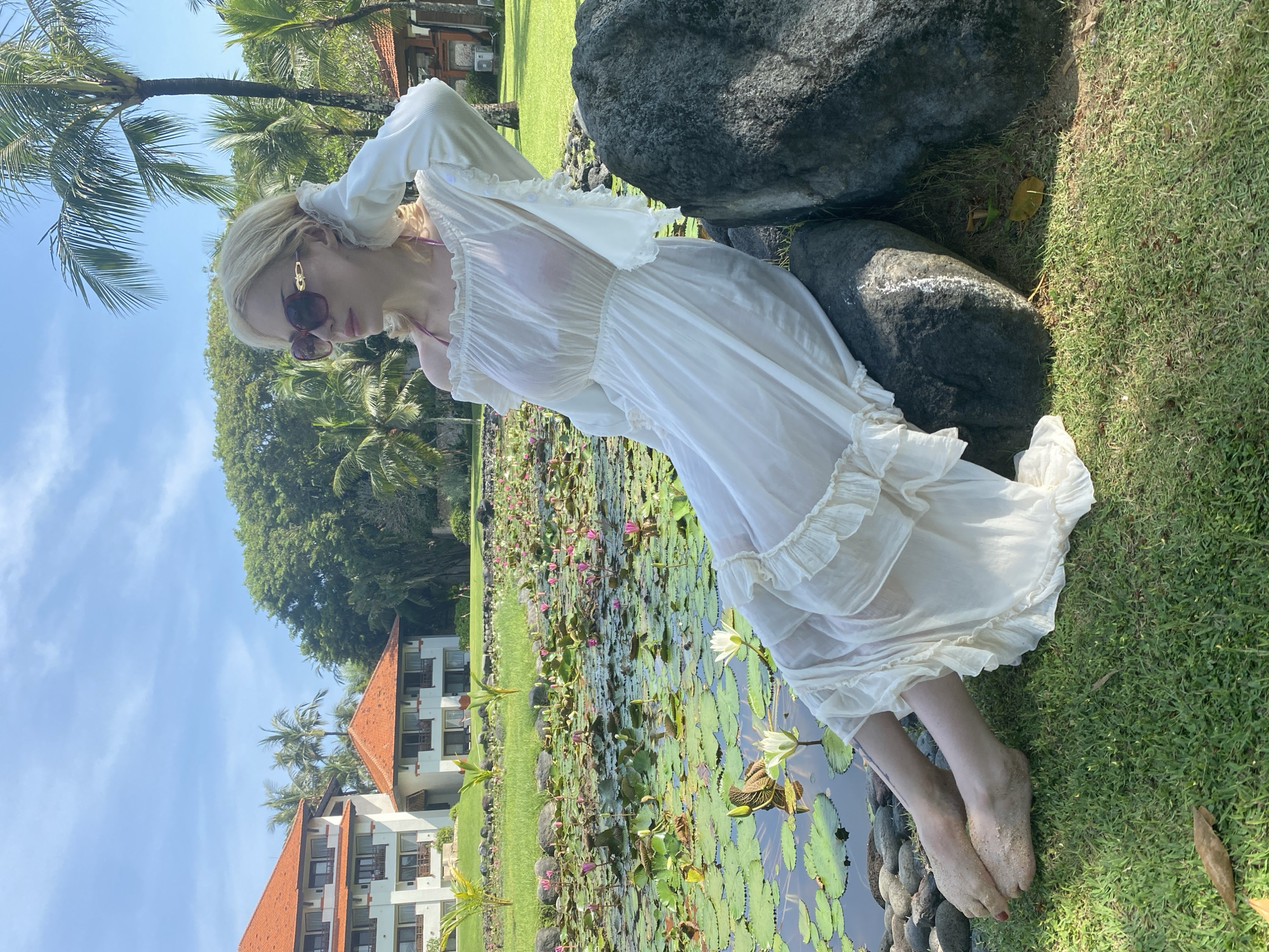 The image depicts a blonde woman in sunglasses and a sheer white dress, leaning against two dark gray rocks on a grassy bank in Nusa Dua, Indonesia. Her bare feet are covered in sand. Behind her, a pond is filled with water lilies. In the background, there is a building with an orange tile roof and palm trees, with a partially visible tree behind it. The sky is blue.