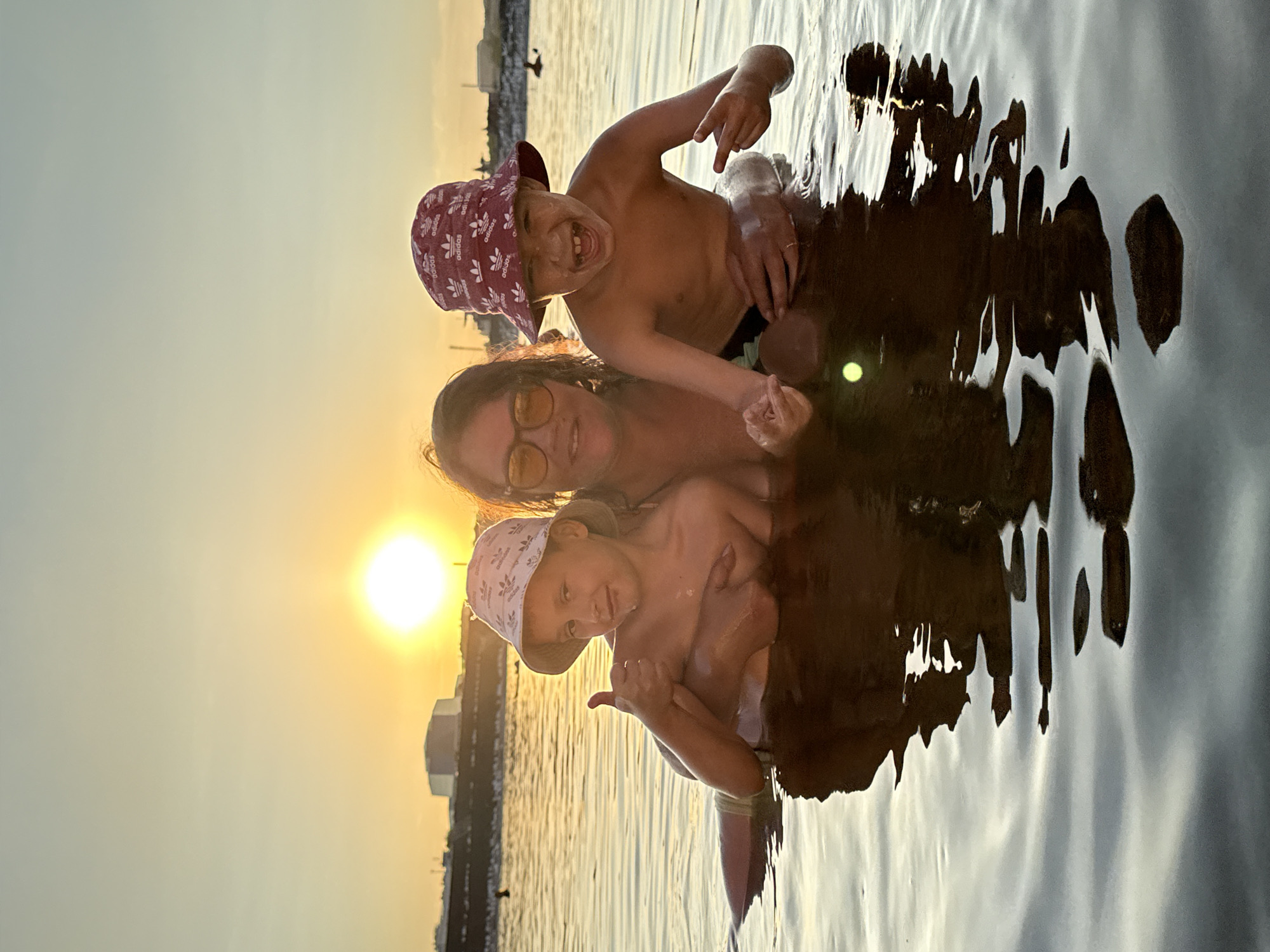 A medium shot captures a family in the water during sunset in Sol-Iletsk, Russia. The sun is low in the sky, casting a warm glow on the water and a distant shoreline with industrial structures. A woman, wearing sunglasses and a dark top, is in the center, embracing two young boys. The boy on the left, wearing a light-colored bucket hat, gives a thumbs-up. The boy on the right, wearing a red bucket hat, grins widely with his mouth open and makes a peace sign with his fingers. Both boys are shirtless. The water is calm with reflections of the sky and the people.