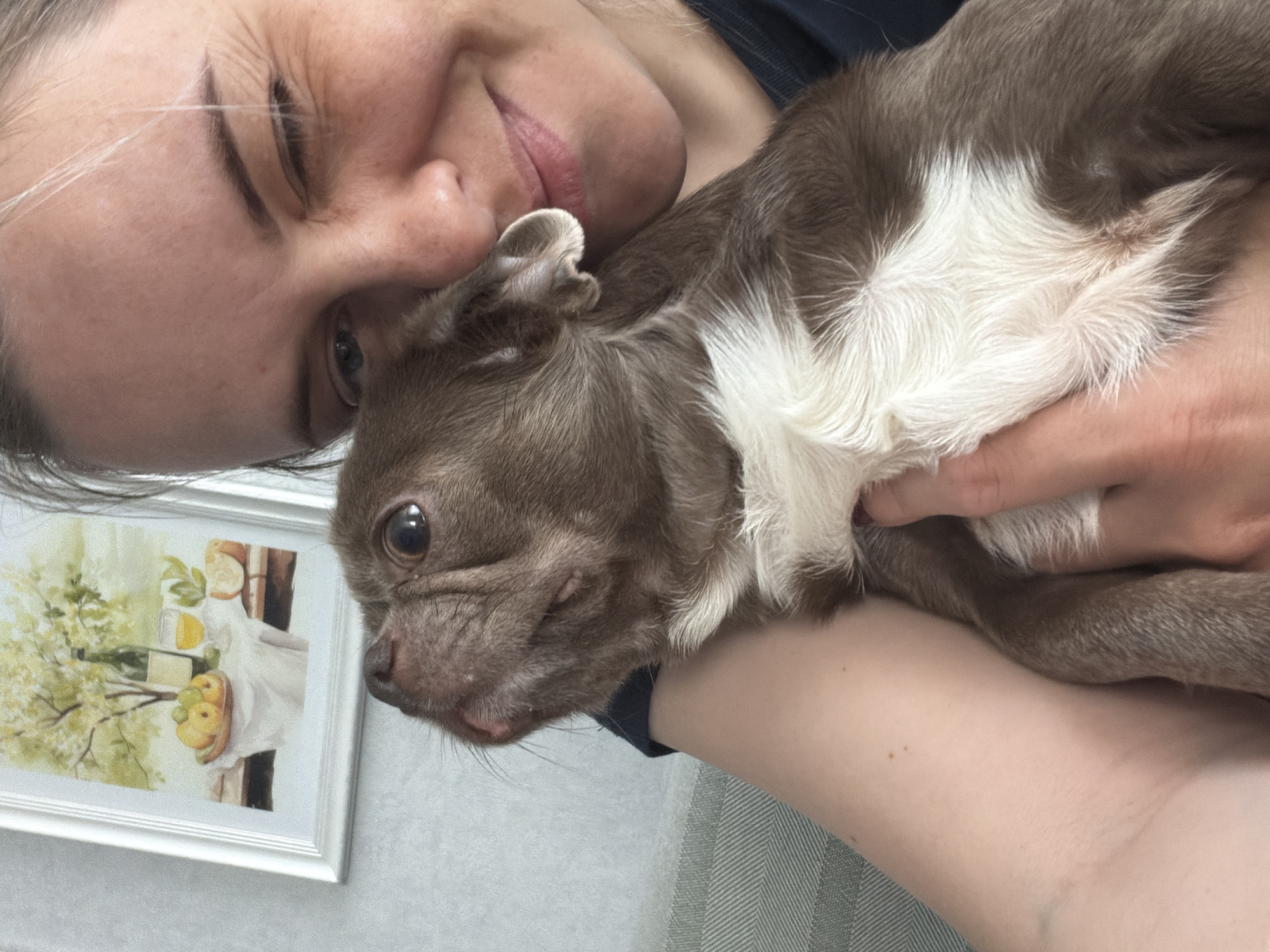 A woman is holding a small brown and white dog. The woman is fair-skinned with blonde hair and is smiling with one eye closed. The dog is brown with a white chest and muzzle. The background includes a framed painting of a still life. The location in the city of Votkinsk, Russia, is not discernible from the image.