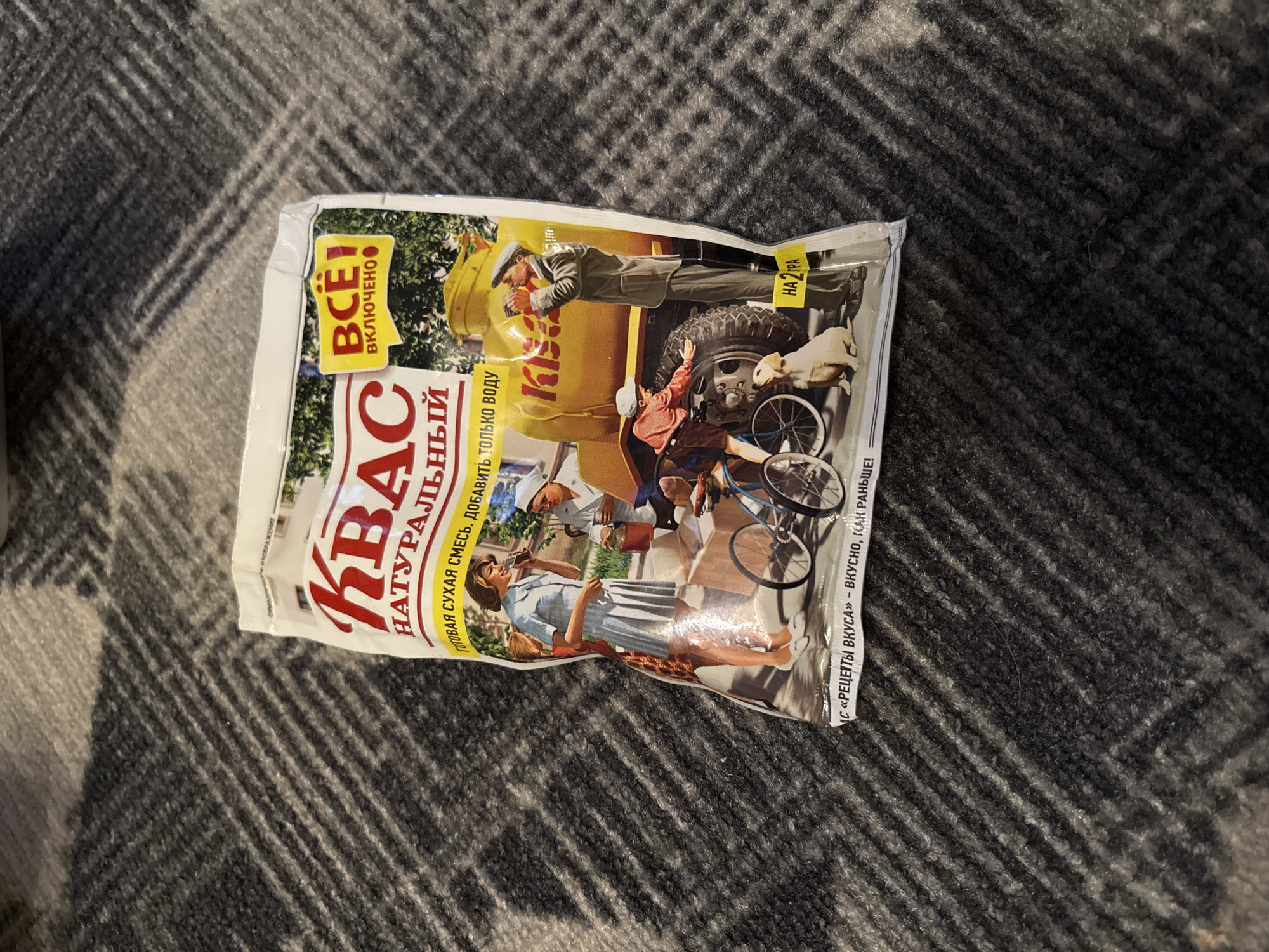 The image shows a close-up, slightly angled shot of a white and yellow package of kvass mix, set against a dark gray, textured surface that appears to be a blanket. The package features vintage-style illustrations.
The front of the package displays the word "КВАС" (KVAS) in large red letters. Below this, in smaller red letters, is "НАТУРАЛЬНЫЙ" (NATURAL). A yellow banner runs across the top, containing the text "ВСТ" (ALL) and "ВКЛЮЧЕНО" (INCLUDED) in black text. Beneath the main title, there is a line of text in a smaller font that reads "ГОТОВАЯ СУХАЯ СМЕСЬ. ДОБАВИТЬ ТОЛЬКО ВОДУ" (READY DRY MIX. JUST ADD WATER).
The central illustration depicts several figures. On the left, a woman in a light blue dress is holding a glass to her lips, seemingly drinking. Next to her, a person in a white hat and uniform is attending to a large yellow tank truck, holding a jar. In front of the truck, a young boy on a bicycle is reaching towards a golden-brown dog that is sitting on the ground. A man in a military-style uniform and cap is standing near the truck, looking towards the boy and the dog, and holding a bottle. The truck has the word "Кіѕ" (KIS) written on its side.
At the bottom of the package, a yellow strip contains the text "НА 2 Л" (FOR 2 L) and further to the left, text that appears to read "«РЕЦЕПТЫ ВКУСА» - ВКУСНО, КАК РАНЬШЕ!" (“RECIPES OF TASTE” - DELICIOUS, LIKE BEFORE!).
The image does not contain any visual cues that would allow for its specific location in the city of Chernogorsk, Russia, or any other specific urban environment. The background is a plain, textured fabric.