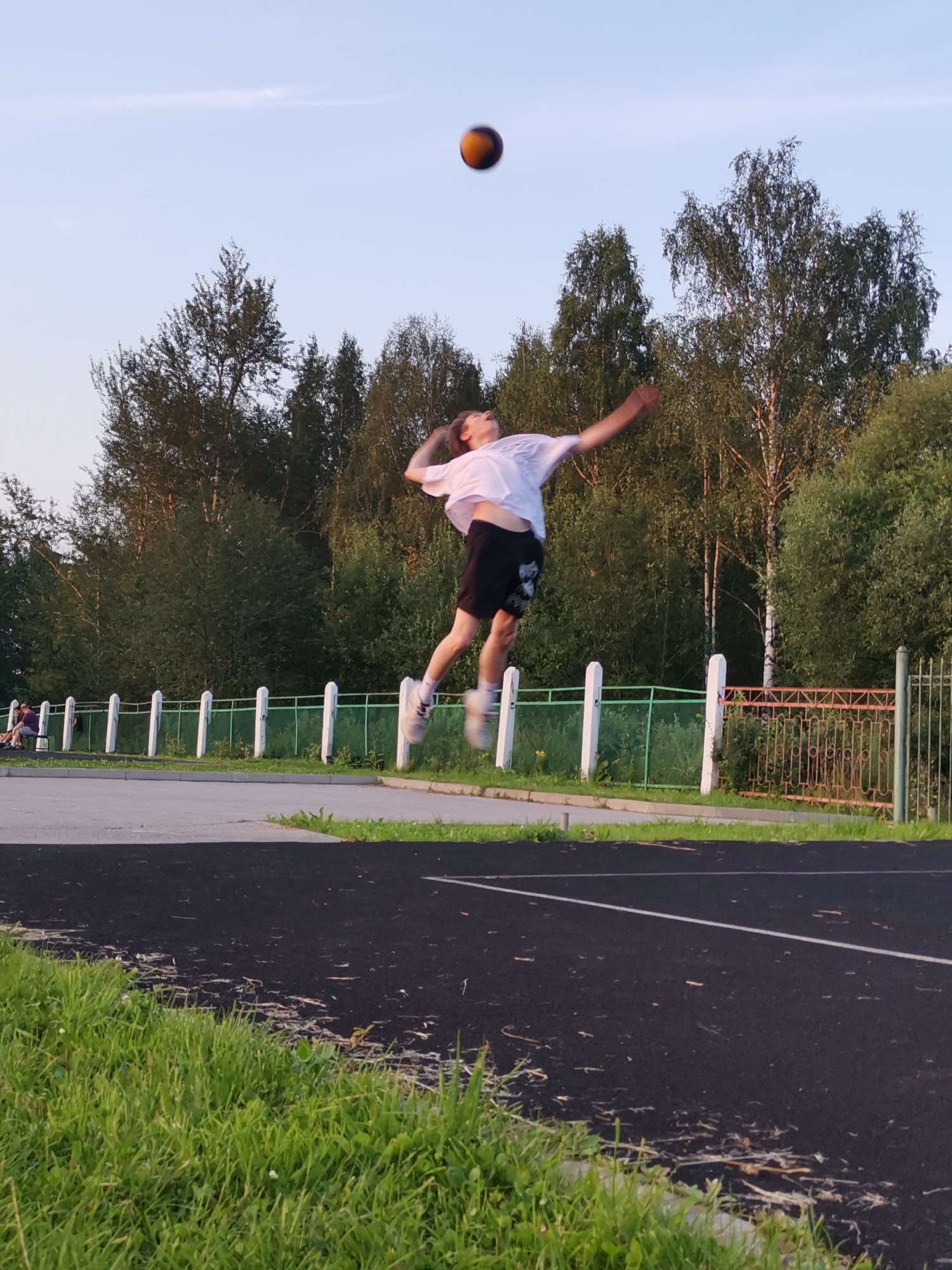 A young male, wearing a white t-shirt and black shorts, is captured mid-air in a volleyball serve. He is positioned in the center of the frame, with the ball just above his head, seemingly having been tossed for the serve. The background consists of a lush green line of trees, a white fence with green netting, and a chain-link fence with an orange and brown pattern. The foreground is a dark asphalt surface with grass on the left. The sky is clear and pale blue. The scene appears to be an outdoor sports area, possibly a park or school grounds in Kizel, Russia.