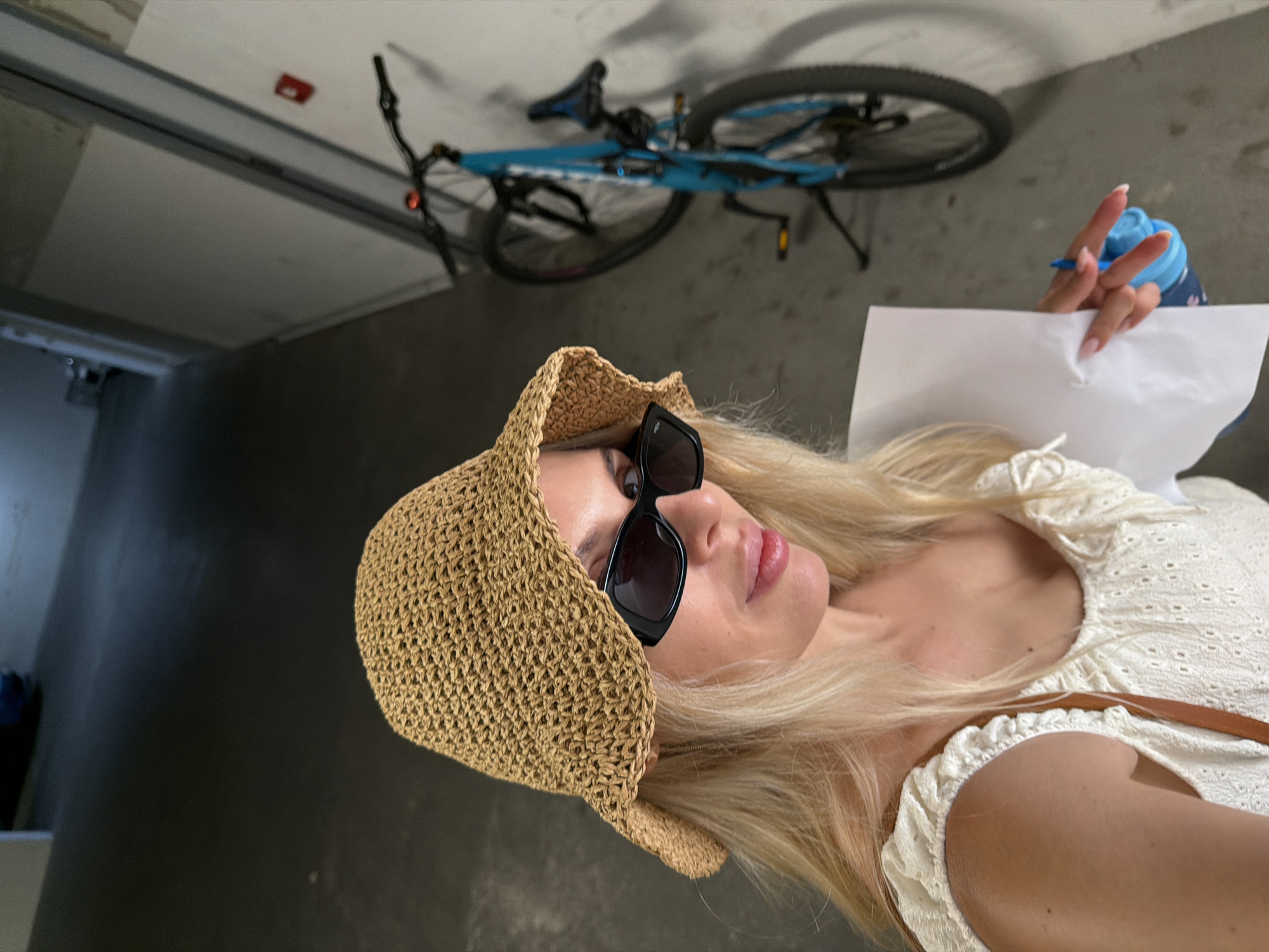 In an indoor hallway or garage, a blonde woman with fair skin, wearing a straw sun hat and black-framed sunglasses, is captured in a mid-shot. She has on a white eyelet top and is holding a piece of white paper and a blue water bottle with a straw in her right hand. Her lips are slightly parted and appear to have lip gloss. In the background, a blue bicycle is propped against a light-colored wall. The floor is concrete and there's a hint of a darker, possibly black, surface in the foreground, suggesting the scene might be in a parking area. The overall lighting is dim, with a spotlight effect illuminating the woman. The location is identified as Budva, Montenegro, within a modern building structure.