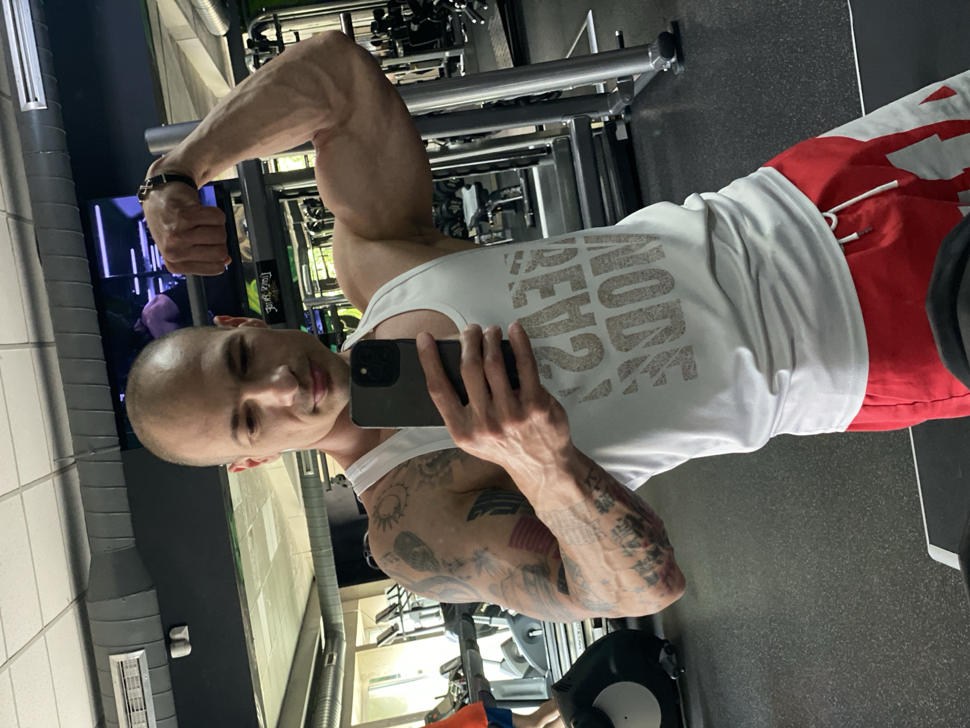 The image shows a bald man in a gym taking a selfie. He is wearing a white tank top and red athletic shorts. His left arm is flexed, showing a developed bicep. The man has multiple tattoos on his right arm. The gym has workout equipment in the background, including weight machines and dumbbells. The location is likely within a gym in Pleven, Bulgaria, as indicated by the prompt.