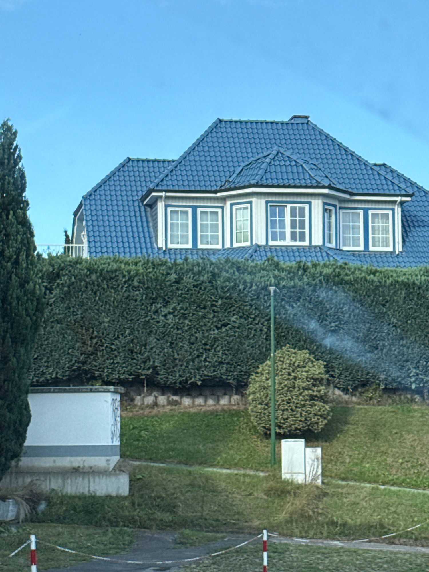 The image shows a blue-tiled roof of a house, with a row of white-framed windows below. A thick, green hedge runs along the front of the property. There is a tall, dark green tree on the left side of the image, and a smaller, round, green bush to the right of the hedge. A thin, green pole stands in front of the bush, and a wisp of smoke is visible in the air near the top of the bush and pole. To the left of the hedge, a white rectangular structure with a grey band around its middle is visible, with some graffiti on its side. The ground in front of the hedge is covered in grass. In the foreground, a red and white striped marker post is visible, with a white and red striped rope extending from it. The scene appears to be a residential area in Osterode am Harz, Germany.