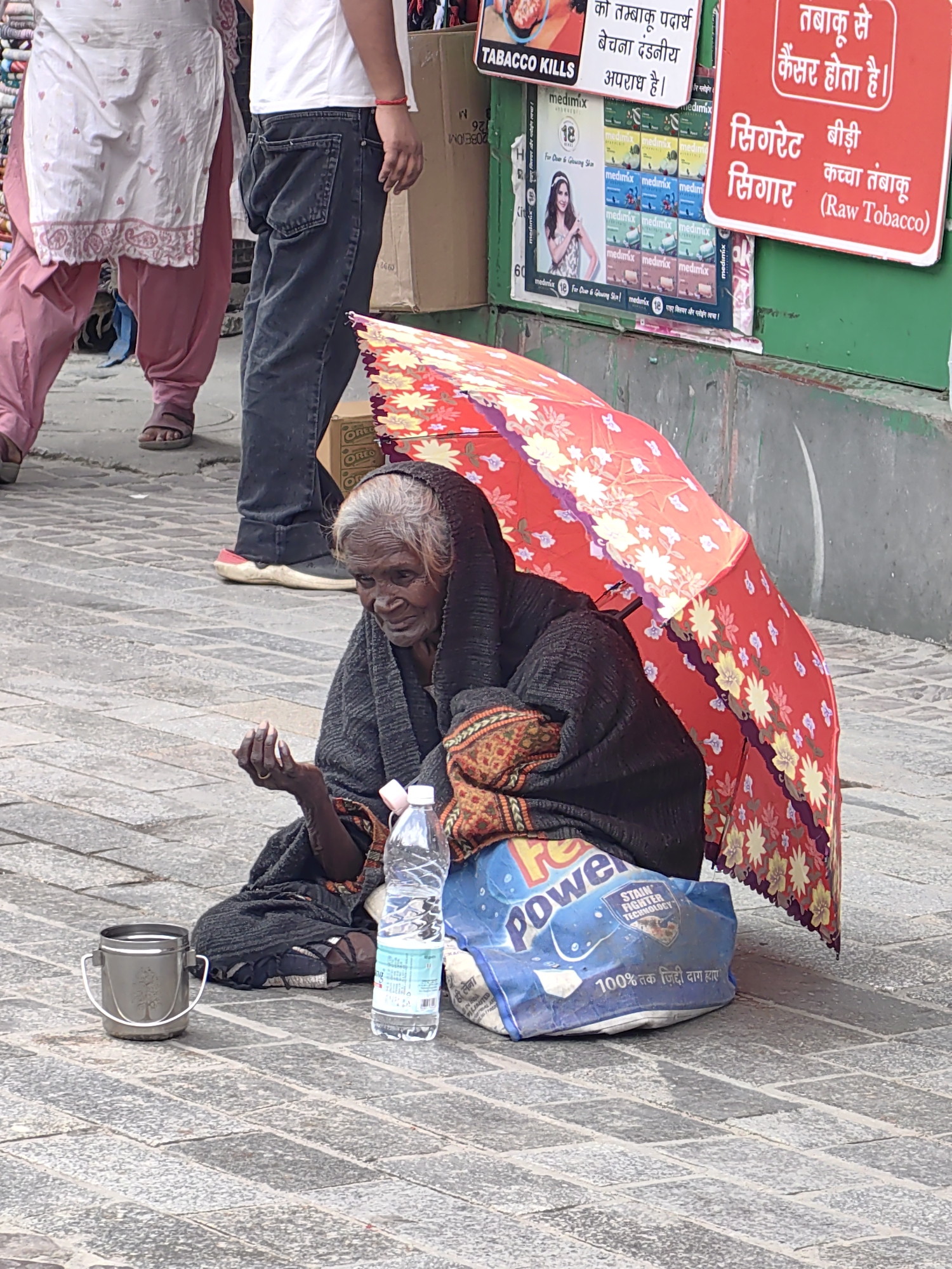 An elderly woman, appearing to be a beggar, is seated on a stone-paved street in Manali, India, partially shaded by a bright red, floral-patterned umbrella. She is wearing a dark shawl and has a bottle of water and a metal container next to her. In the background, a person wearing a white t-shirt and jeans walks past, along with other individuals. The scene includes advertisements on a green wall, suggesting a commercial area.