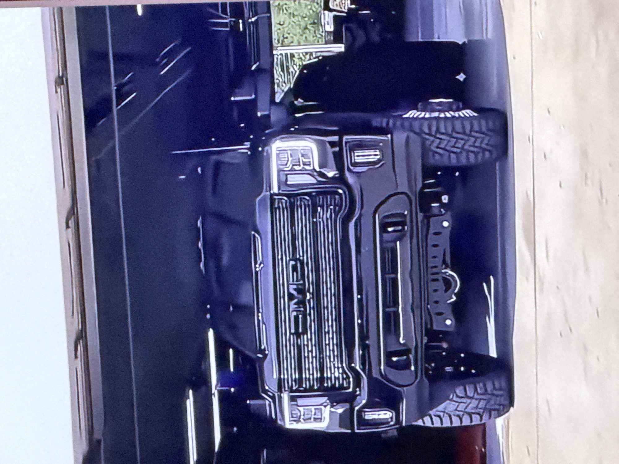 A black GMC Sierra truck is parked inside a garage. The truck is viewed from the front and is the primary subject of the image. The truck has large, aggressive tires and a chrome grille with the GMC logo. The headlights are illuminated. The garage appears to be a workshop or service bay, with overhead lights visible and some equipment in the background. The floor of the garage is concrete. The background shows a window with greenery visible outside. There are no people visible in the image. The location is not specified as Cahul, Moldova.
