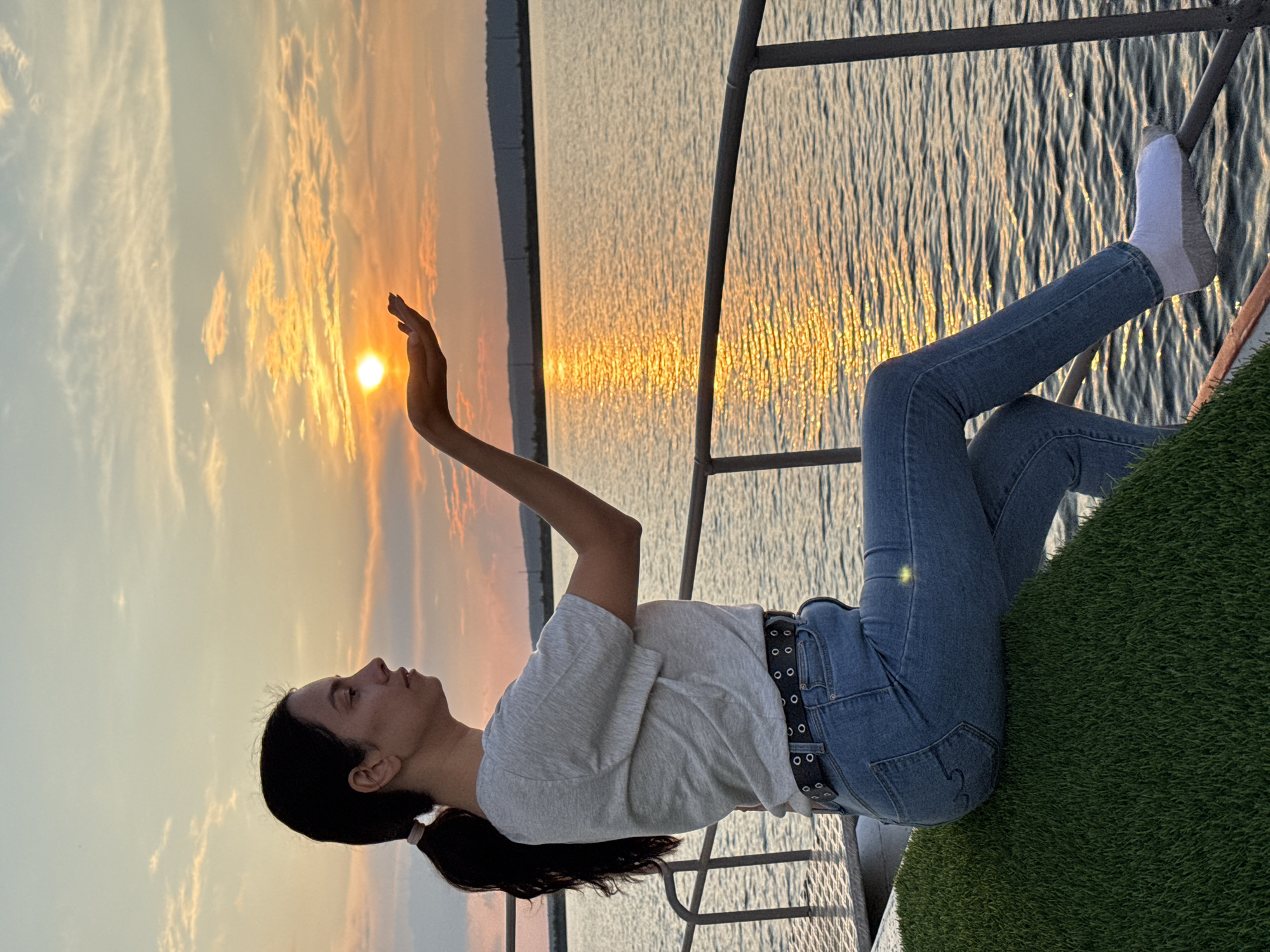 A woman is sitting on a green surface, possibly artificial turf, next to a railing overlooking a body of water. The woman has dark hair tied in a ponytail and is wearing a grey t-shirt, blue jeans, and white socks. She is holding her right hand out, cupping it to seemingly capture the sun, which is setting over the water, casting a golden glow. The sky is filled with soft clouds in hues of orange and yellow. The water reflects the sunset, creating a shimmering path of light. In the background, a distant shoreline with hills is visible. The scene suggests a peaceful moment at sunset on a waterfront, possibly a promenade or park area in Chaikovsky, Russia, given the presence of a large body of water and the characteristic landscape.