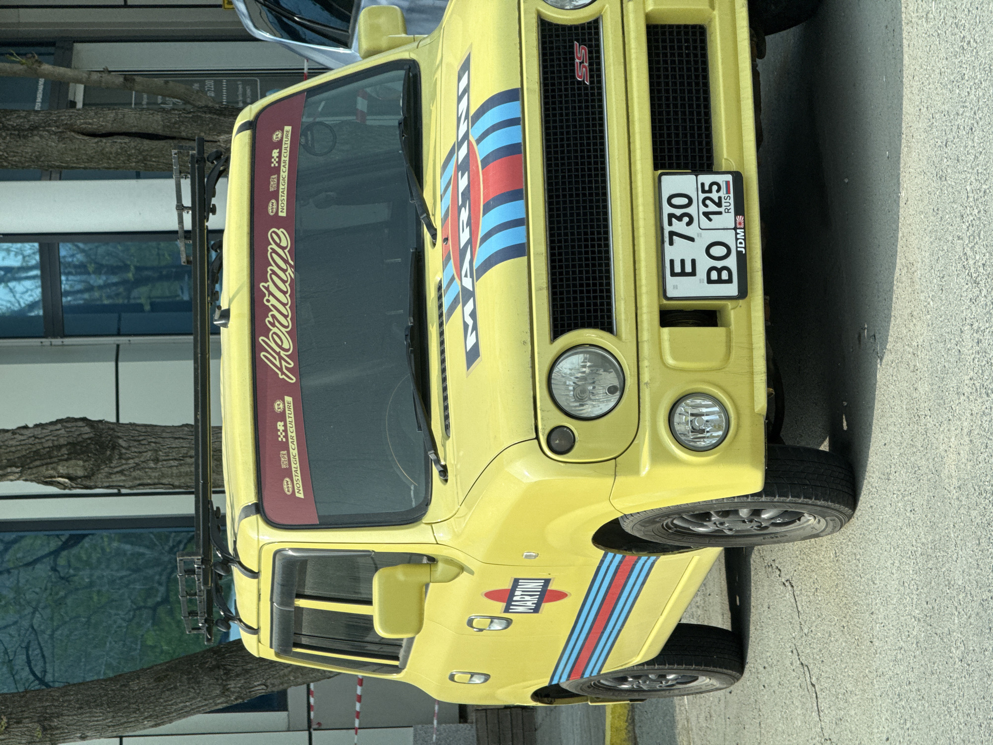 The image shows a yellow Suzuki Jimny car with Martini racing stripes parked in Vladivostok, Russia. The car has a roof rack and decals on the windshield saying "Heritage NOSTALGIC CAR CULTURE." The license plate reads "E 730 BO 125" with a "RUS" designation and a "JDM" label. The grill has an "SS" badge. The car is parked next to a tree and a building with reflective windows. The building has an advertisement stating "до 22:00".