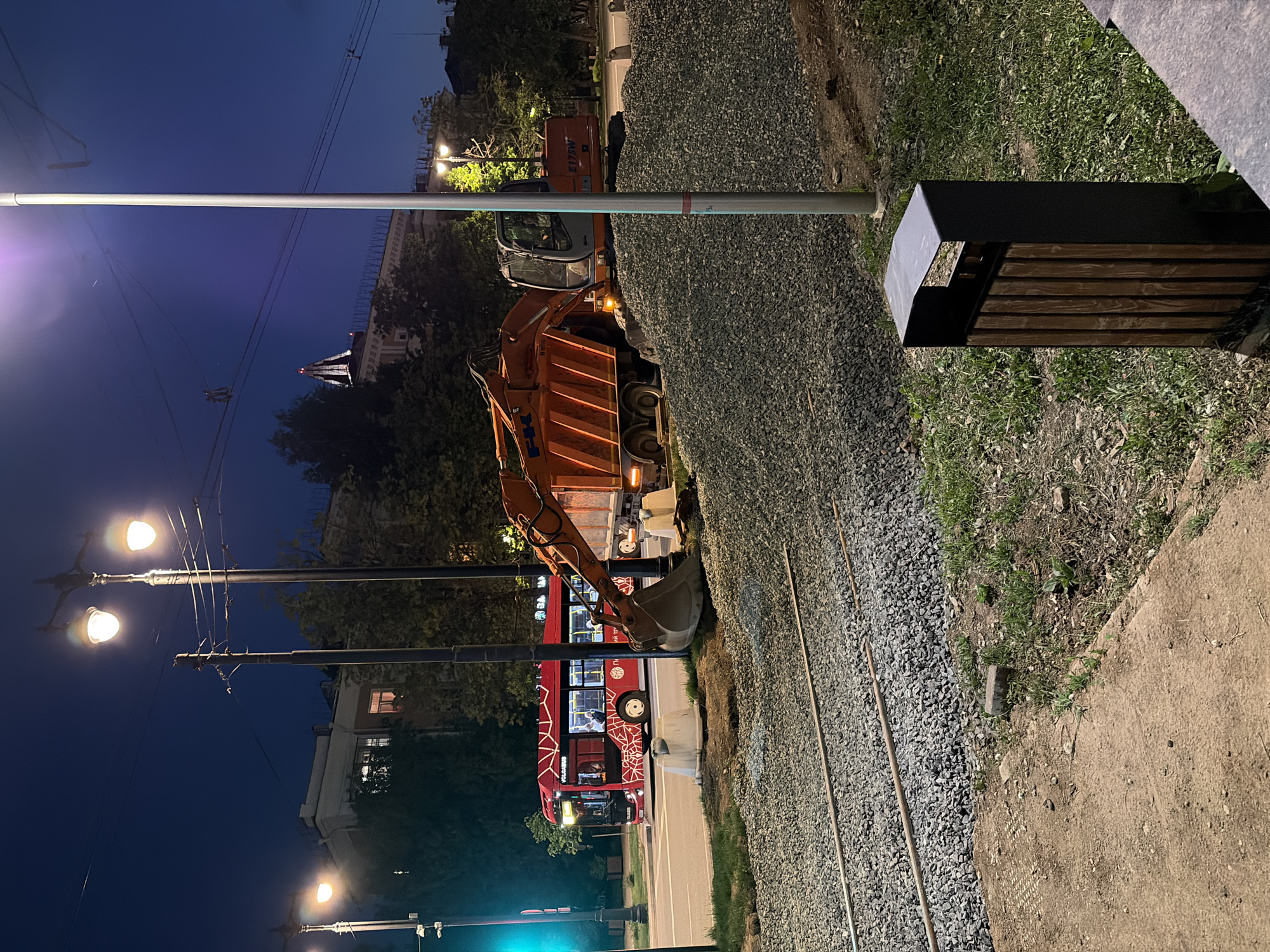 The image depicts an outdoor scene at night in Perm, Russia, near the intersection of Bazisnyy Street and Ulitsa Revolyutsii. A red "CityZen" bus is visible on Ulitsa Revolyutsii. An excavator, potentially part of an infrastructure project, is adjacent to the bus and a pile of gravel, suggesting ongoing construction or maintenance along the street and railway line. A street lamp illuminates the area, with a modern-style trash can/bench located to the side of the street. Overhead electrical wires are apparent. The sky is a deep blue, typical of nighttime. The image shows street repairs.