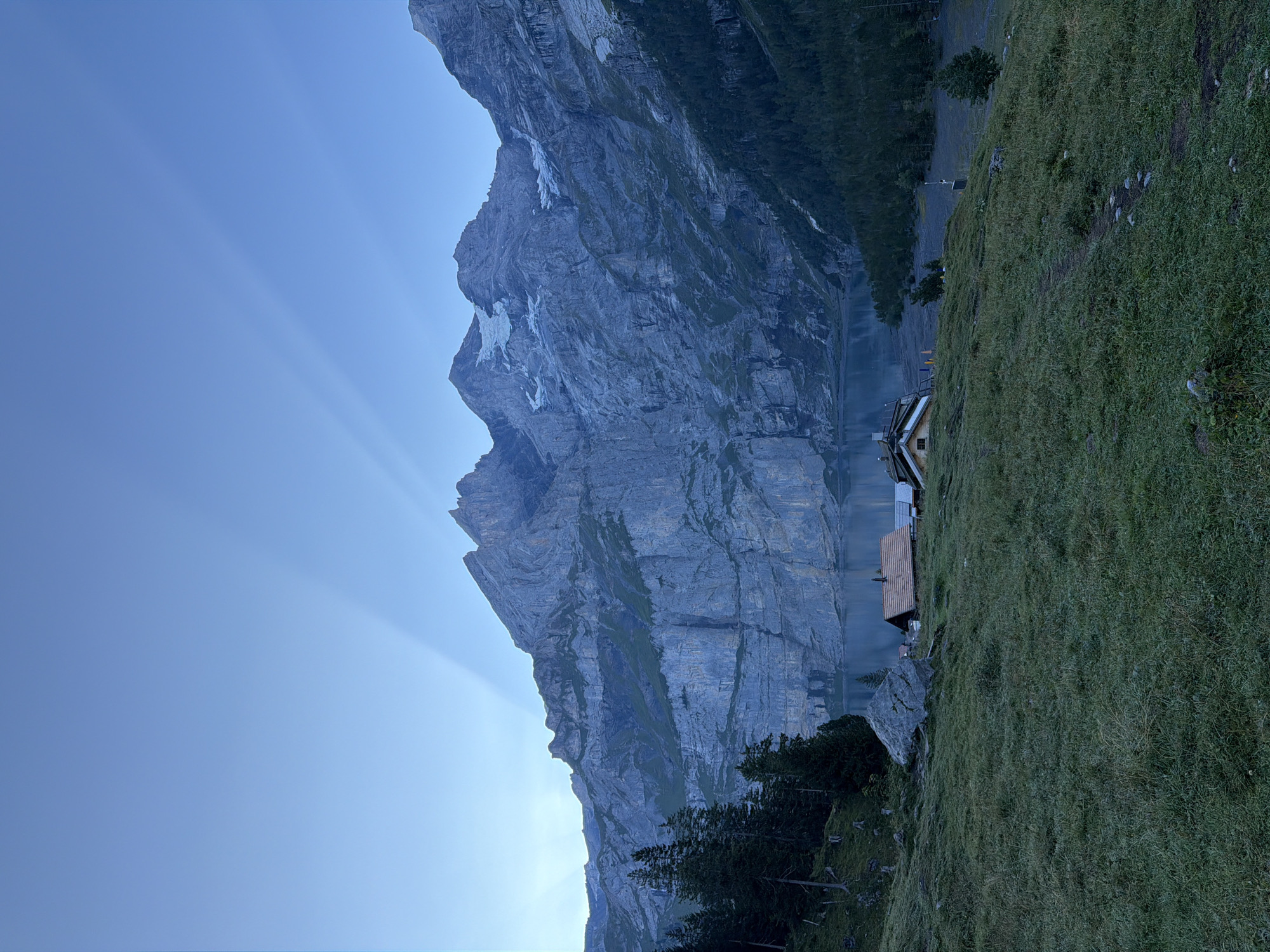The image depicts a serene mountain landscape at Oeschinensee, near Kandersteg, Switzerland. In the foreground, a gently sloping grassy meadow is dotted with a few small rocks and a winding dirt path. A large boulder is situated to the left of the meadow. Two rustic wooden buildings with orange roofs are nestled on the hillside, overlooking a calm lake. Beyond the lake, towering, rugged mountains with sheer rock faces rise dramatically. Patches of snow are visible on the higher peaks. The sky above is a clear, pale blue, with distinct sunbeams fanning out from behind the mountain peaks, suggesting either sunrise or sunset. Dark green pine trees line the edges of the meadow and the lake. The overall impression is one of tranquility and natural beauty, characteristic of the Swiss Alps.