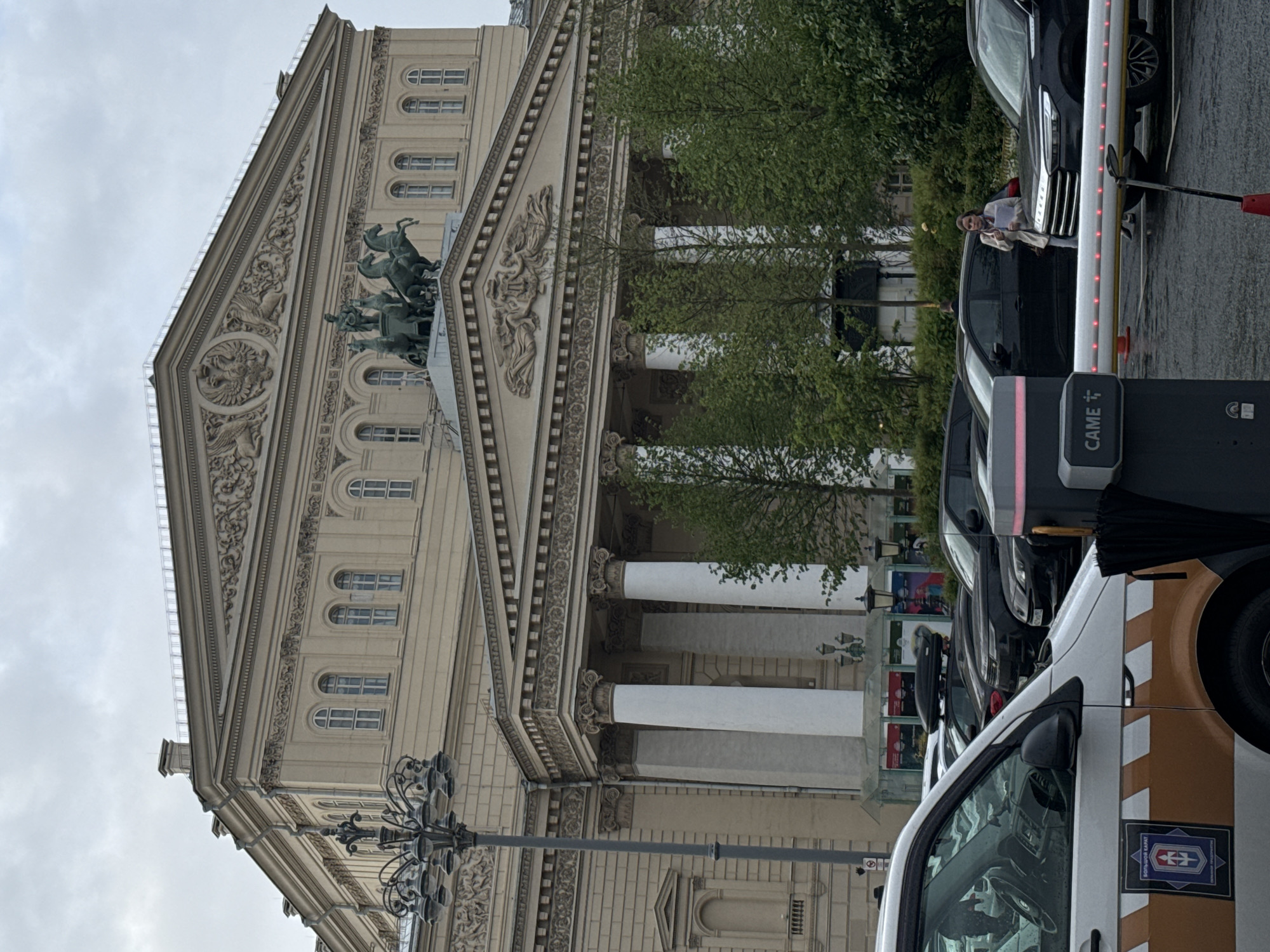 A long shot shows the Bolshoi Theatre in Moscow, Russia, adjacent to a street filled with parked vehicles.
The pale-colored theatre building features a classical architectural design. A sculpture of a horse-drawn chariot adorns its facade. Tall, white columns form a colonnade along the front of the building.
The street alongside the theater is occupied by a line of parked cars. A "CAME" branded gate or barrier, with red LED lights, is visible near the front of the line of vehicles. A woman stands beside one of the dark-colored cars, holding what appears to be a document. A white van, with brown and white stripes and a black logo, is parked in the foreground.
The sky is overcast and a bit gray. Trees are growing behind the line of parked vehicles.