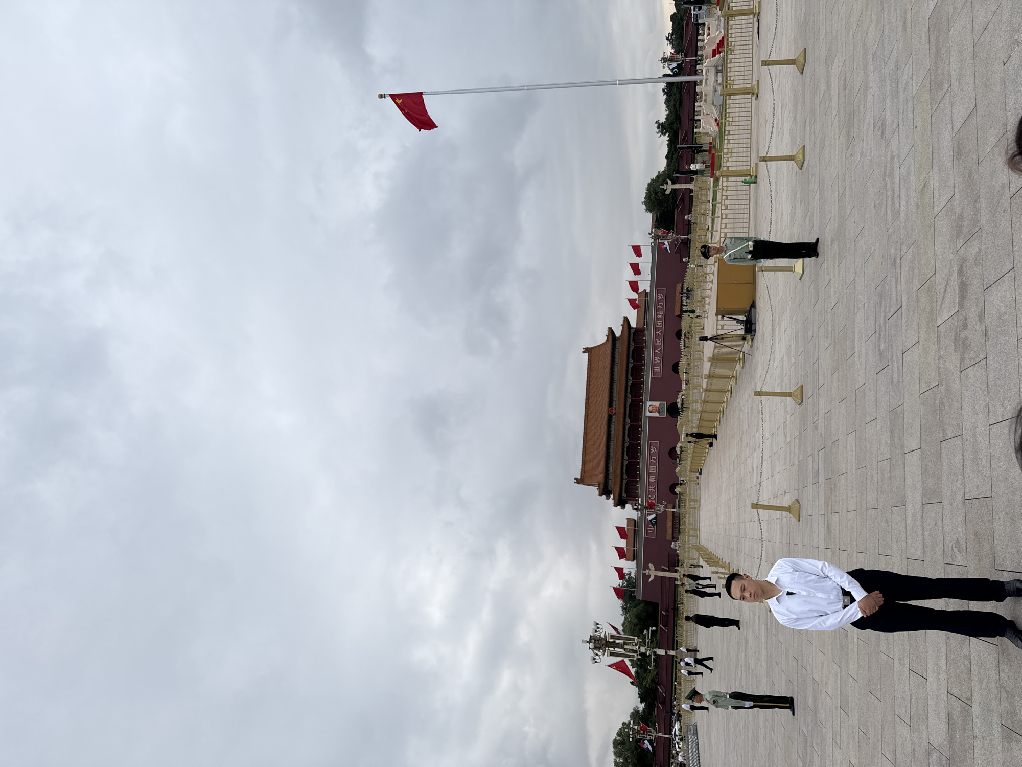 The image captures a scene in Tiananmen Square, Dongcheng District, China. In the foreground, a man in a white shirt and black pants is standing. Behind him, guards stand watch. A flagpole flies the Chinese flag. The Tiananmen Gate is visible in the background, adorned with red banners and a portrait. The sky is overcast. The square is paved with gray stone. Security barriers are erected along the edge of the square.
