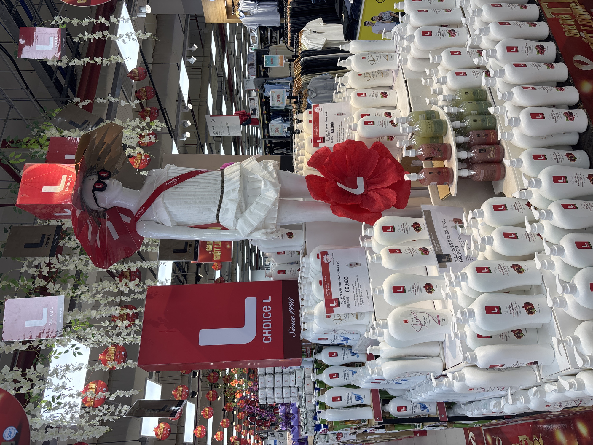 The image shows a display of "Choice L" brand body wash in a store, likely in Đà Nẵng, Vietnam. Rows of white bottles with red "L" logos are stacked on shelves. A mannequin wearing a white dress and sunglasses is positioned near the display, adorned with a large red flower and the "Choice L" logo. Signage indicates promotional pricing for the body wash. There are clothing racks with shirts in the background. The store appears decorated with floral garlands and hanging lanterns. Some signs are written in Vietnamese.