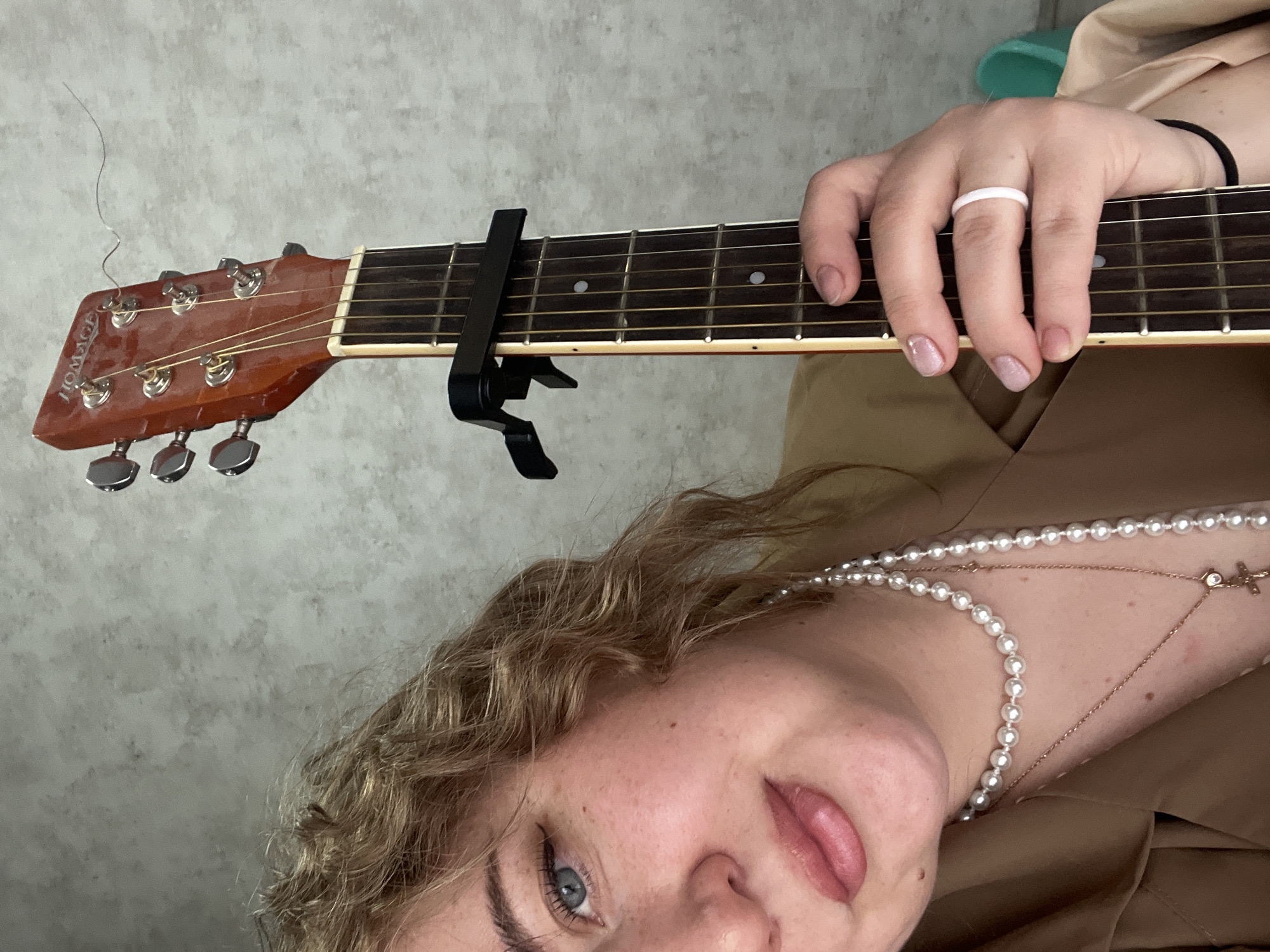 The image displays a young woman holding an acoustic guitar, captured from a slightly overhead perspective with the guitar neck extending horizontally across the upper half of the frame. The woman's face, featuring her right eye, nose, and mouth, is visible in the lower right quadrant, turned towards the viewer. She has light blue eyes and curly blonde hair framing her face. Her lips are slightly parted, and her tongue is subtly visible, conveying a relaxed or playful expression. She wears a brown or beige top.
Around her neck are two necklaces: one composed of small white pearls, and another thinner, gold-colored chain with a tiny cross pendant. Her right hand rests on the guitar's fretboard, fingers gently curved, with a plain white ring on her ring finger and a black hair tie on her wrist. Her nails have a light, shimmery pink polish.
The guitar, a "HOMAGE" brand, features a reddish-brown headstock with silver tuning pegs. A thin, wavy metal string is detached and extends upwards from the headstock. A black capo is clamped onto the fretboard, positioned after the first fret. The fretboard itself is dark with white dot markers. The background is a plain, light gray textured wall, suggesting an indoor setting. There are no specific time of day or weather cues visible.
