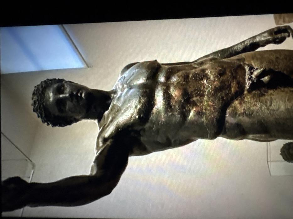 A bronze statue of a muscular, nude male figure is depicted in an indoor environment, likely a museum or gallery. The statue, aged with a greenish-brown patina and reflective highlights, is positioned at an angle, with its right arm raised above its head and its left arm partially obscured, hanging downward. The figure features short, curly hair and an idealized classical facial structure, with visible genitalia. The background consists of a plain, light-colored wall. A rectangular blue light source, possibly a window or skylight, is visible in the upper right. In the lower left, a dark, partially visible object, resembling another statue's head or bust, is enclosed within a transparent display case. The image does not contain any visual identifiers to confirm the specific location as Jos, Nigeria.