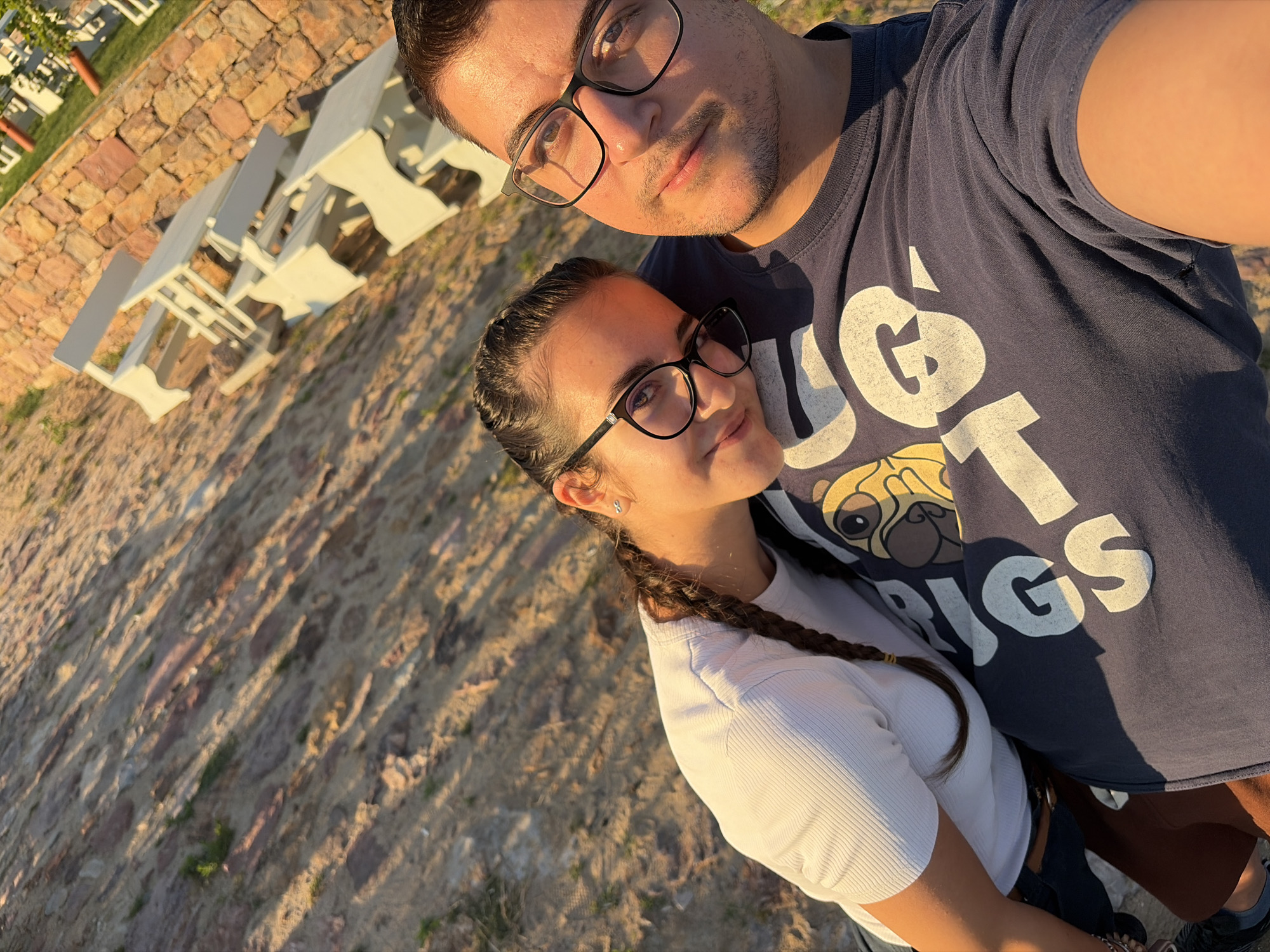 In Sofia, Bulgaria, a man and a woman are taking a selfie outdoors, possibly at a picnic area. The woman, with dark hair styled in a braid and wearing glasses and a white top, is resting her head on the man's shoulder. The man, also wearing glasses and a navy t-shirt with a pug graphic and white lettering, is looking directly at the camera. The background features a stone wall, a sandy ground, and several white picnic tables. The scene is illuminated by warm, late-afternoon sunlight, casting shadows across the ground and highlighting the textures of the stone and sand.