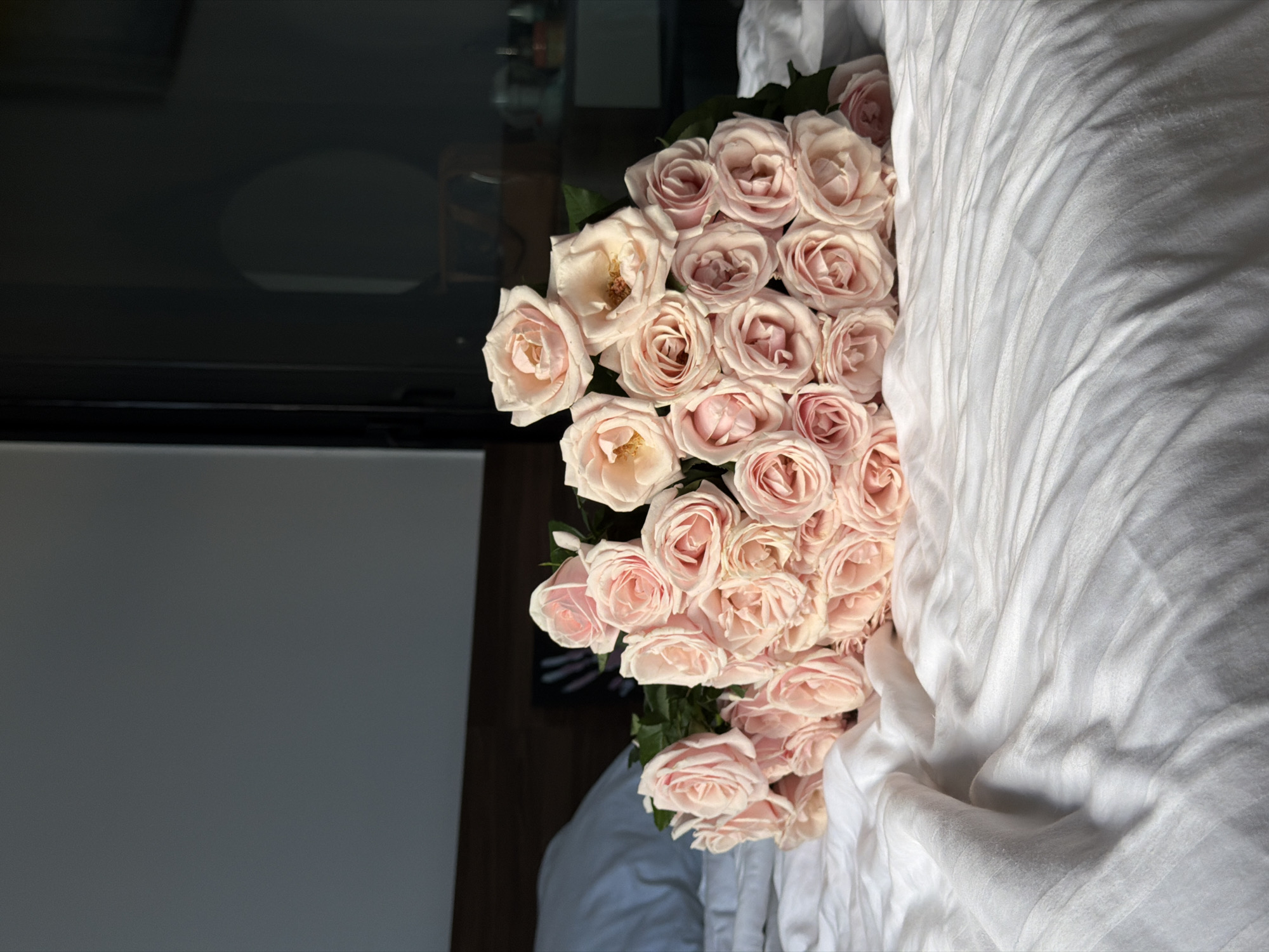A bouquet of light pink roses is arranged on a rumpled white bedspread. The bed is positioned in what appears to be a bedroom, with a dark wooden headboard visible behind the roses and a pillow on the left side of the frame. The room is dimly lit, with light entering from the right, casting shadows and highlighting the texture of the bedsheets and the petals of the roses. The background on the right is dark and reflective, suggesting a window or glass door. There are no people visible in the image. This scene is not specific to any location in Da Nang, Vietnam, as there are no identifiable landmarks or contextual clues.