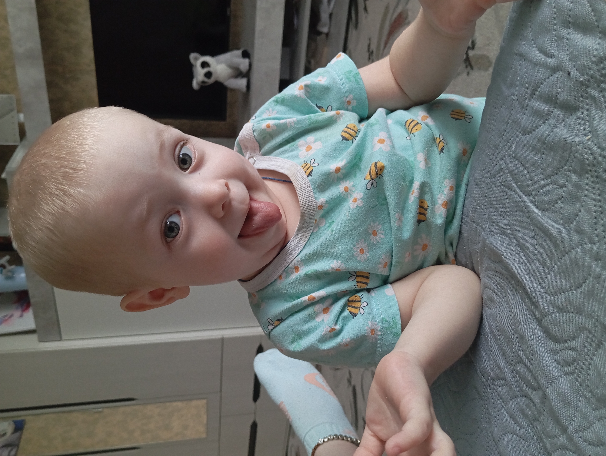 A young child with blonde hair and light skin is sticking out their tongue. The child is wearing a light green shirt with white daisy and bee patterns. The child is on a quilted, light blue surface, presumably a bed or blanket. A small, gray stuffed animal is visible on a shelf in the background. A sock with a pink logo is partially visible in the lower left corner of the image. The location is likely indoors in Yurgamysh, Russia, but this cannot be confirmed from the image alone.
