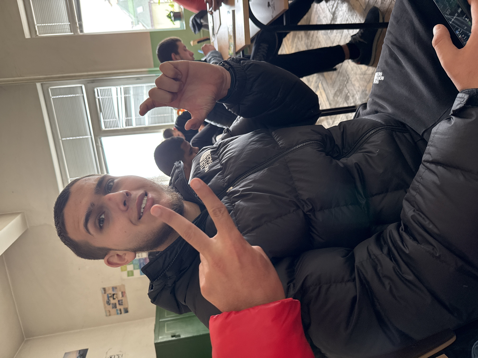 A young man is posing indoors, likely in a classroom setting, with several other people present in the background. He's wearing a black puffer jacket with a red sleeve accent. He is gesturing with both hands, making a "peace" sign with one hand and extending his thumb and pinky finger with the other. The setting appears to be a school classroom, as indicated by the windows, blinds, and the arrangement of desks. The exact location within Sofia, Bulgaria, cannot be determined from the image alone, as there are no specific landmarks or signs visible.