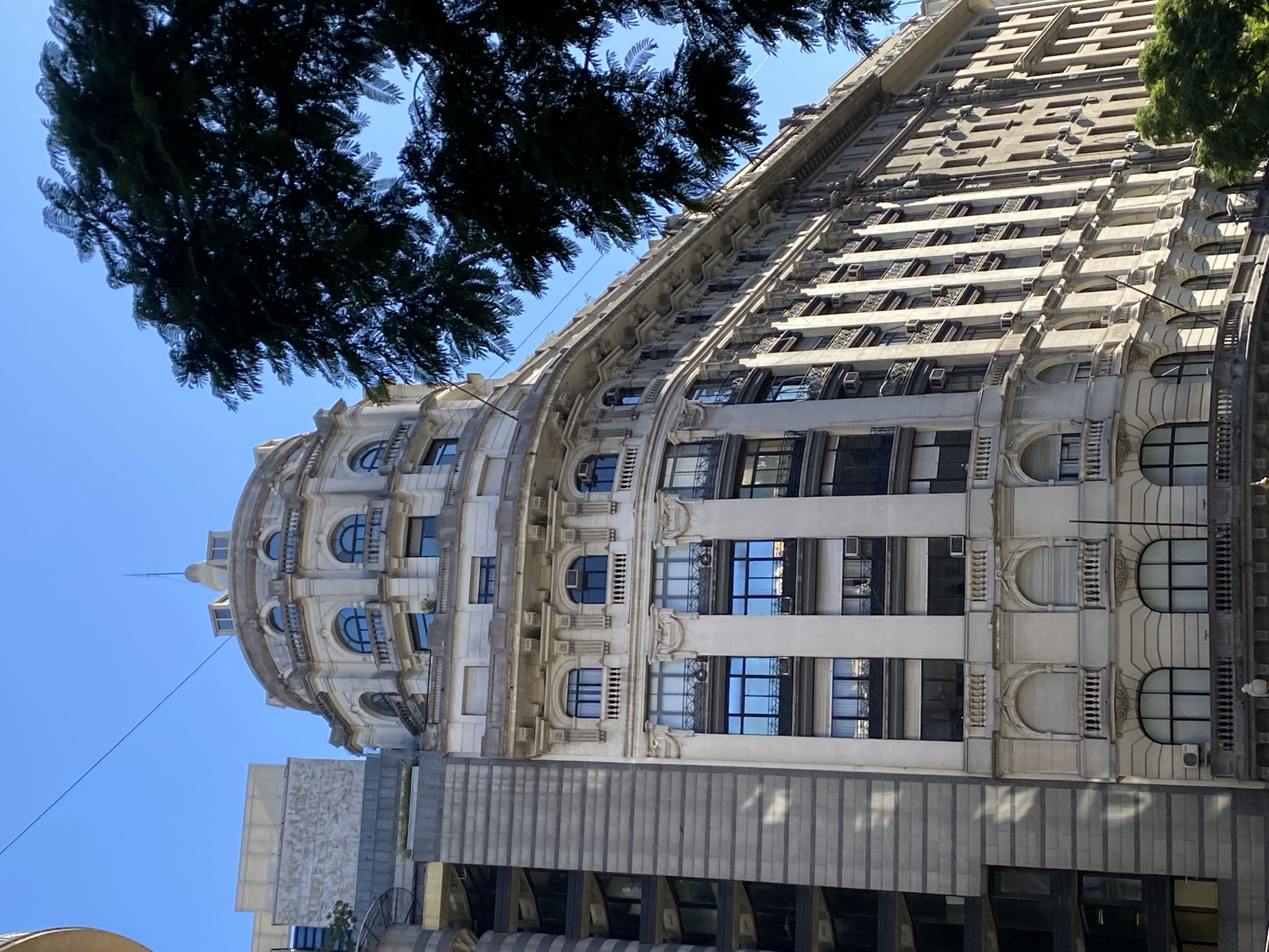 An upward shot captures the historic Bencich building in Buenos Aires, Argentina, from a low angle. The building's facade is ornate, featuring rounded arches, columns, and decorative elements typical of early 20th-century architecture. The upper floors are marked by numerous windows reflecting the bright blue sky. Air conditioning units are visible in some windows, indicating modernization. The top of the building is crowned with a small dome or cupola. A tree with dark green foliage partially obscures the top left corner of the frame. At the building's base is a more modern structure with a minimalist gray facade. The lighting suggests a sunny day.