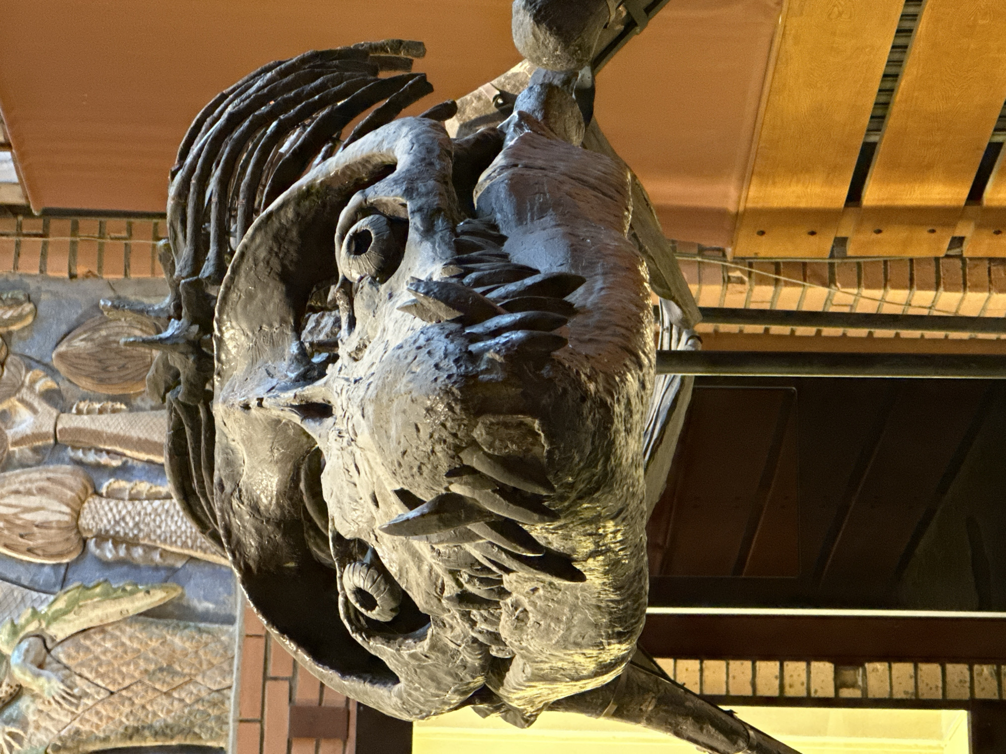 The image depicts a large, dark-colored sculpture, possibly made of metal or stone, resembling the head of a prehistoric creature with prominent teeth and eye sockets. The sculpture is viewed from a low angle, looking up. The background includes decorative elements such as carved panels featuring depictions of lizards and other objects. Behind the sculpture are architectural details like a ceiling with wooden planks and a brick wall. The location is Moscow, Russia, as it is the only city where the State Darwin Museum is located, which houses this sculpture.