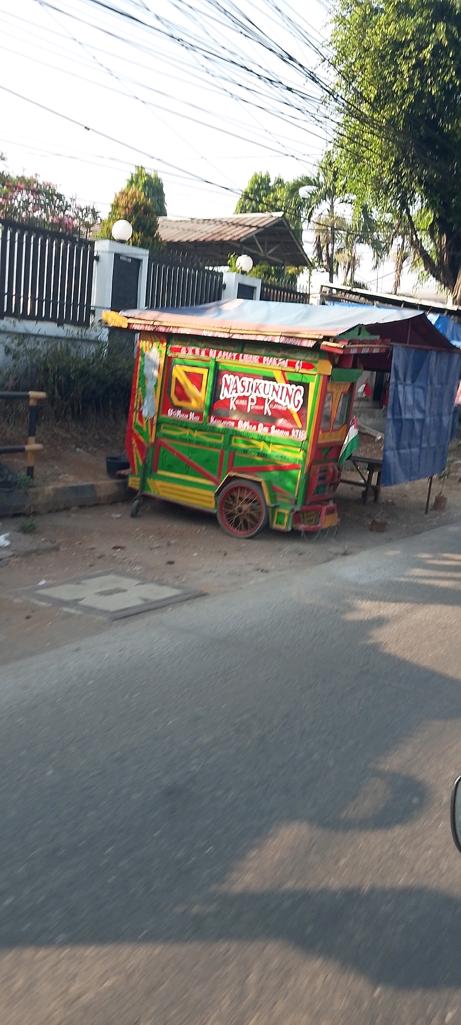 The image depicts a vibrant, colorful food cart, likely a "Nasi Kuning" (yellow rice) vendor, parked on a dirt roadside in Cileungsi, Indonesia. The cart is predominantly green, yellow, and red, with lettering on its side that reads "NASI KUNING". It has a canopy covering and a single wheel visible at the rear. In the background, a black metal fence with white posts and a house with a tiled roof are visible, suggesting a residential or commercial area. The ground is a mix of dirt and pavement, with a manhole cover present on the pavement. Overhead, a network of power lines crisscrosses the sky, and a large tree with dense foliage shades part of the scene. The lighting suggests it is daytime, possibly late afternoon, with shadows cast across the road. The scene is static, with no people or activity explicitly shown.