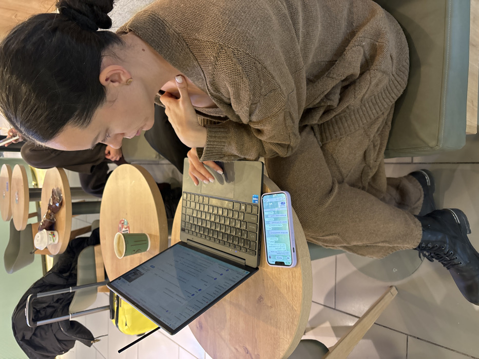 The image depicts an indoor scene, identified as a cafe or waiting area, in Novogorelovo, Russia. In the foreground, a woman, appearing to be in her late 20s or early 30s, is seated at a round wooden table. She wears a brown cable-knit cardigan over matching brown trousers and black lace-up boots with silver accents. Her dark hair is pulled back into a bun. Her left hand is positioned on the touchpad of an open silver laptop, which displays a list-based interface. Her right hand rests near her collarbone. A light purple-cased smartphone, displaying a messaging application with multiple chat bubbles, lies face-up on the table next to the laptop. A green paper coffee cup is also on the table, behind the laptop. In the background, another round wooden table is visible, where a second individual, partially obscured and wearing dark clothing, is seated. A white coffee cup and a brown wrapper are on this table. To the far left, a black rolling luggage bag with a black jacket draped over it is present, next to a yellow bag. The floor is light-colored tile, and seating throughout the area incorporates green upholstered elements.