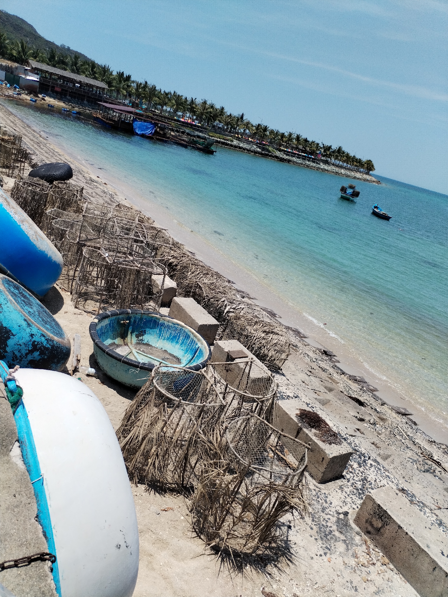 The image depicts a coastal scene in Bac Nha Trang Ward, Vietnam. In the foreground, there are several round boats and fishing traps situated on a concrete embankment and sandy beach. The boats are in various states of repair, some painted blue and white. The fishing traps are made of netting and dried palm fronds. Concrete blocks are scattered along the embankment.
Behind the boats and traps, the image transitions to a sandy beach with some dried vegetation. The clear turquoise water of the bay is visible. Further out, there are two boats anchored in the water. The coastline is lined with palm trees and some buildings, suggesting a populated area. The sky is blue with minimal cloud cover.