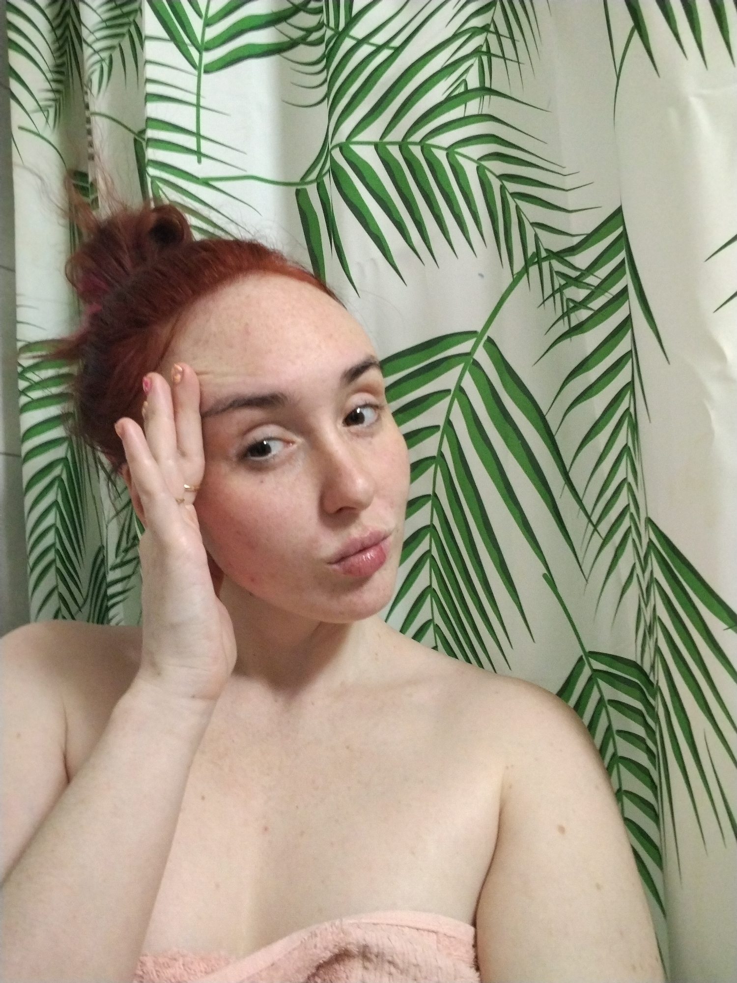 A photograph depicts a close-up, head-and-shoulders view of an adult female in an indoor setting. The specific location within the city of Izhevsk, Russia, cannot be determined from the visual information; however, the background is a white shower curtain patterned with green palm fronds, indicating a bathroom environment.
The female subject has fair skin with visible freckles on her face, shoulders, and upper chest. Her reddish-brown hair is pulled up into an untidy bun. Her eyes are dark and appear to be looking directly at the camera. Her lips are slightly puckered. She is bare-shouldered, with a peach-colored towel draped around her upper body, obscuring the lower chest area. Her right hand (viewer's left) is raised, with fingers splayed and resting against her forehead. A gold-colored ring is visible on the ring finger of her right hand. Her fingernails are painted, with the thumb showing a light color and the ring finger displaying a gold or glitter polish. The lighting is even, suggesting an indoor light source.