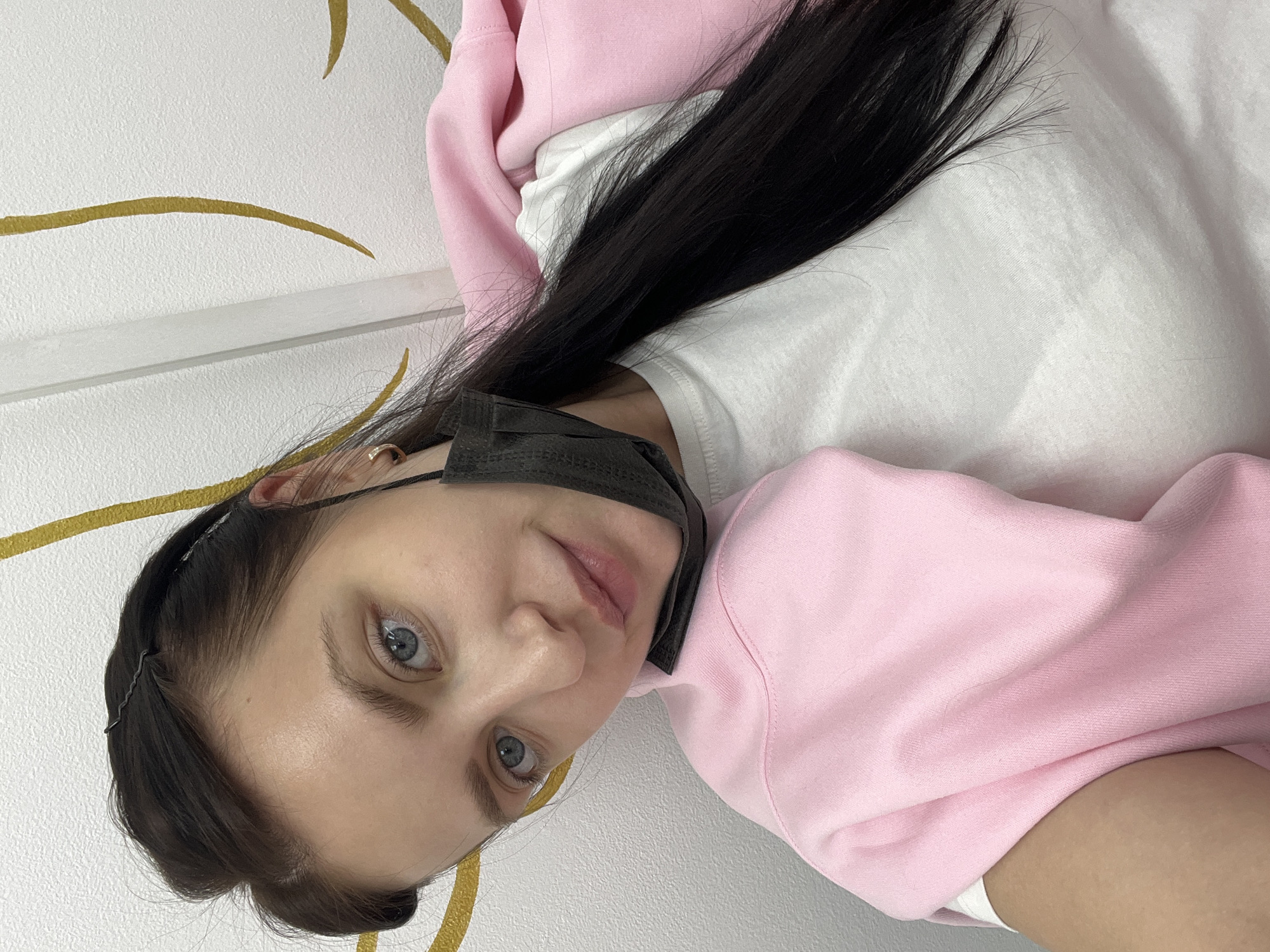 This close-up portrait features a young person, likely female, with fair skin, captivating blue eyes, and long, straight dark hair. They are looking directly into the camera with a calm and neutral expression. The individual is dressed in a light pink hooded sweatshirt, worn over a white t-shirt. A black protective face mask is visible around their neck, pulled down and resting below their chin. Their dark hair, parted on one side, cascades over their shoulder and chest, with a small section near the forehead secured by a black bobby pin or hair clip.
The background consists of a brightly lit, white textured wall, which features delicate, abstract golden wavy lines as decoration. A narrow white horizontal molding or shelf runs across the wall behind the subject. The setting appears to be indoors, with consistent lighting. There is no discernible activity beyond posing for the photograph. The geographical context for this image is Chaikovsky, Russia. No text is visible in the media file.