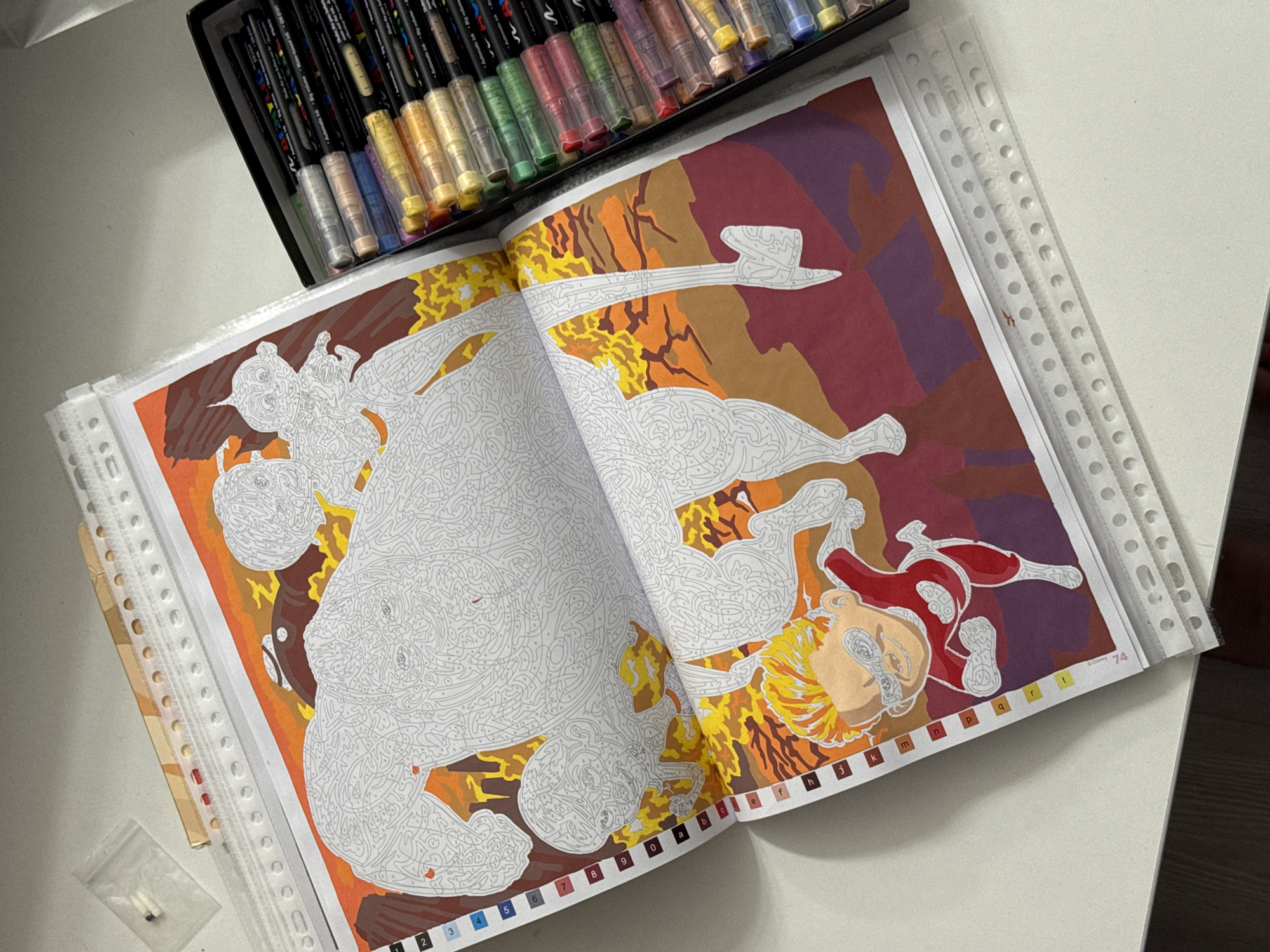 The image shows an open paint-by-numbers book on a white surface, next to a black box containing a set of colorful markers. The paint-by-numbers book depicts a scene with large, abstract figures in shades of white and grey, with a partially colored background in oranges, yellows, reds, and purples. A small figure of a character with blonde hair and a red suit is visible in the lower part of the book, also partially colored. The markers in the box are of various colors, including green, pink, yellow, and blue, and appear to be Posca paint markers. The image is taken from a slightly elevated angle. There is no indication of any location in Łódź, Poland.