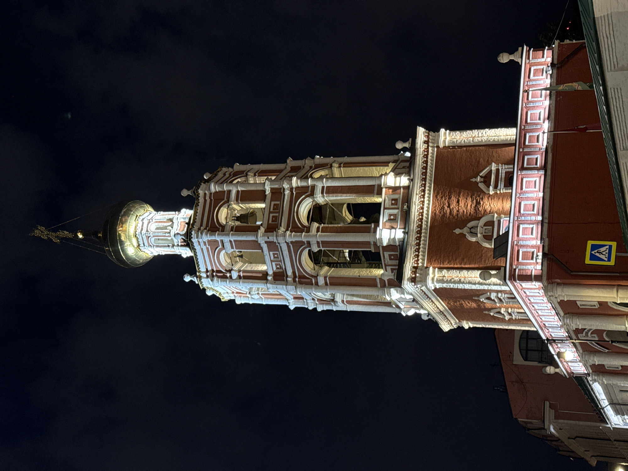 The image shows a close-up, low-angle night shot of the bell tower of the Church of Saints Cosmas and Damian on Maroseyka Street in Moscow, Russia. The bell tower is illuminated by artificial lights, highlighting its red brick structure with white decorative elements. The tower is topped with a golden dome and a cross. In the foreground, a portion of a red building with a white balustrade is visible, along with a yellow pedestrian crossing sign. The background is a dark, cloudy night sky.