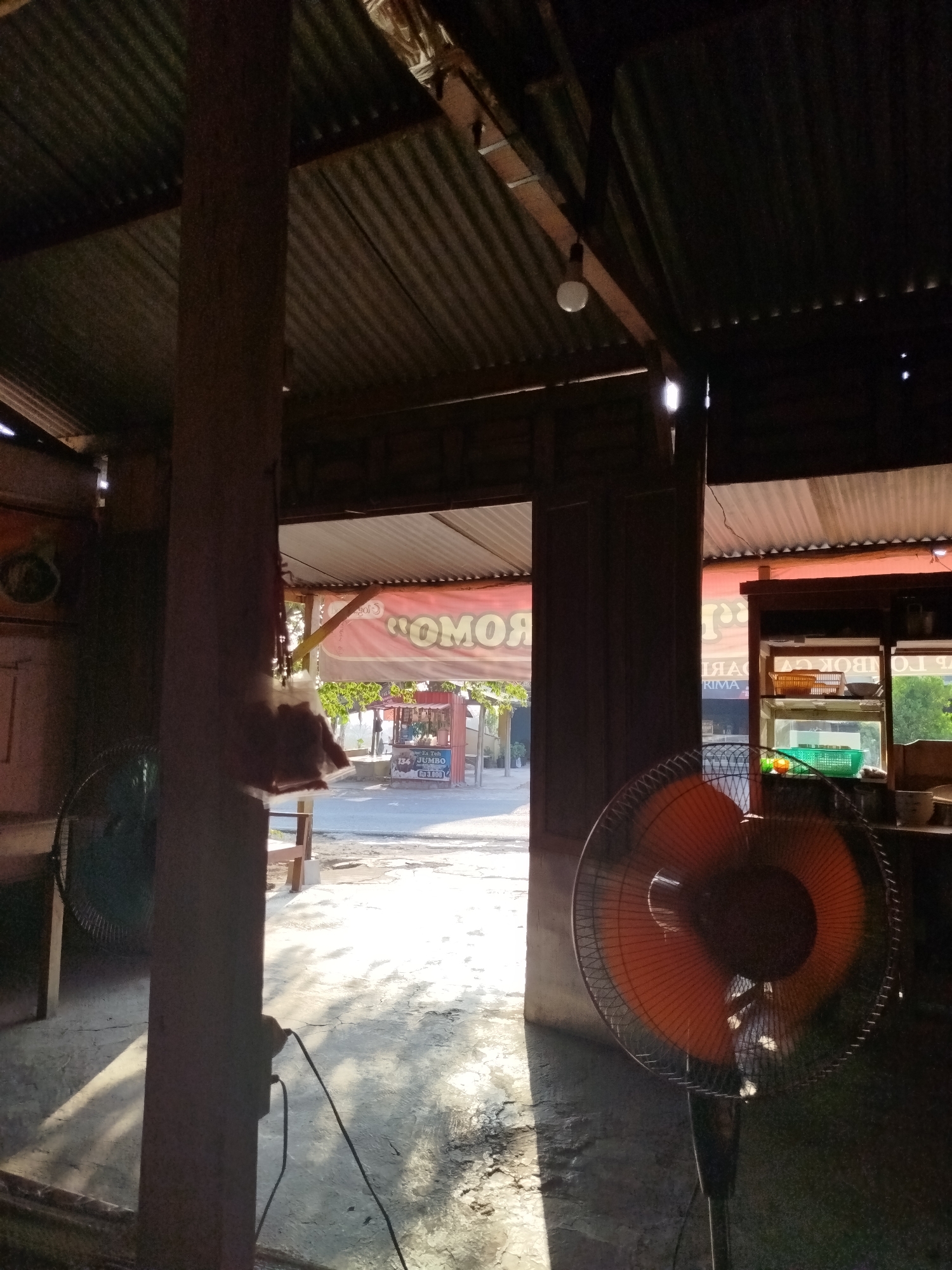 The image depicts the interior of a simple structure, possibly a shop or eatery, with a corrugated metal roof. Sunlight streams in from an opening, casting strong shadows across the concrete floor. In the foreground, two electric fans are visible: one is dark green and partially obscured by a wooden pillar, while the other has bright orange blades and is positioned more centrally. The wooden pillar is a prominent vertical element in the scene.
Beyond the immediate interior, an outdoor area is visible through an open doorway. This area appears to be a street with a light-colored surface. In the distance, a small kiosk or food stall is set up, featuring a red banner with white text that is partially obscured and appears to be a local advertisement. A person can be seen standing near this stall. The environment suggests a warm, sunny day. The specific location in Pojok, Indonesia, is not explicitly indicated by any visible signage, but the general appearance is consistent with a small business in a tropical or sub-tropical region. The overall scene conveys a sense of everyday life in a community setting.