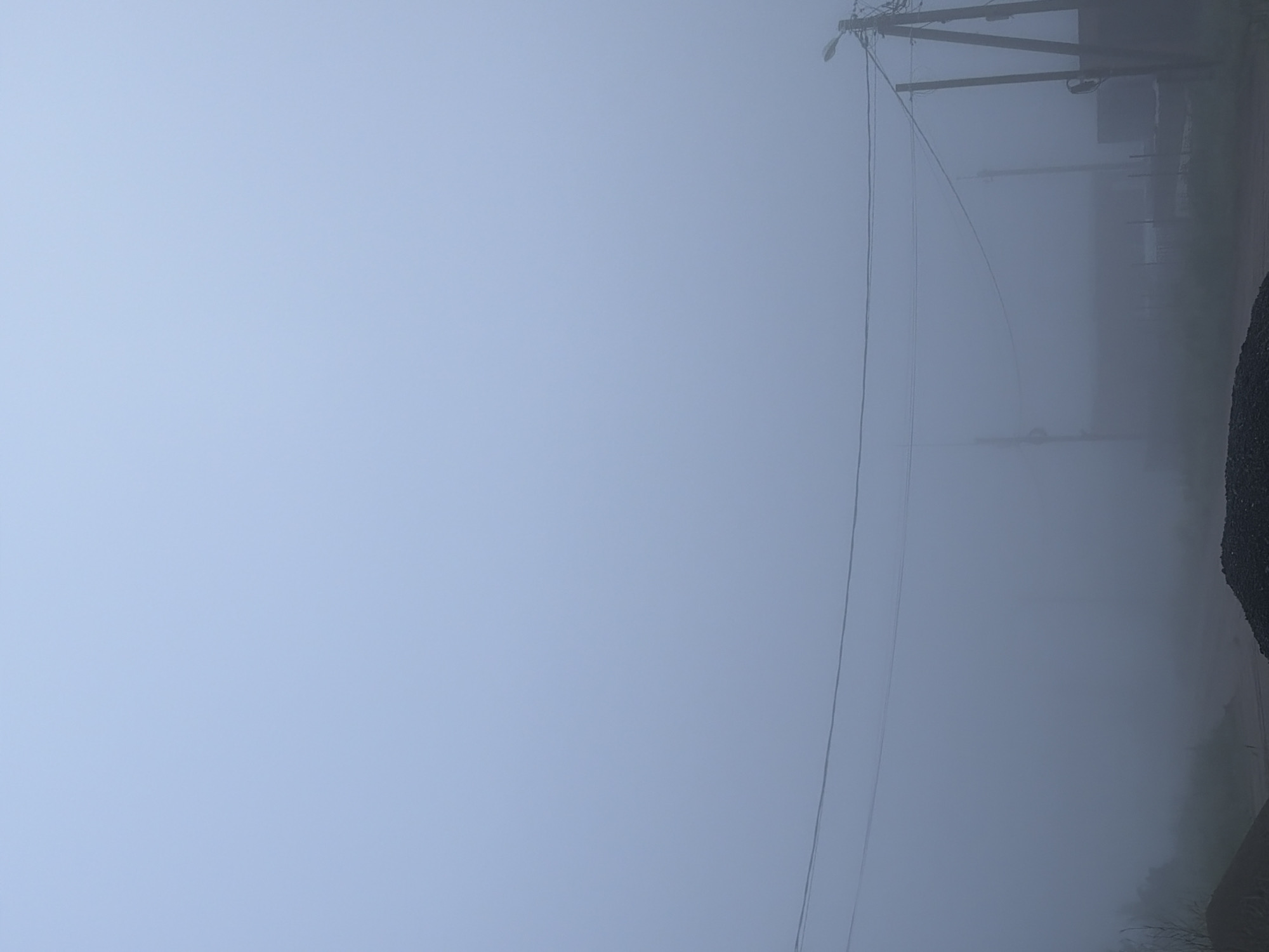 The image, originally oriented vertically, depicts a landscape heavily obscured by dense fog or mist. The predominant color palette is a soft, uniform blue-gray, reflecting the pervasive atmospheric conditions and extremely low visibility.
On the right side of the frame, a dark utility pole, appearing to be made of wood, stands partially visible, its details softened by the mist. A street lamp is attached to the upper part of this pole. Multiple power lines extend horizontally across the mid-ground and upper-right portions of the image, gradually disappearing into the haze. Further into the background on the right, very faint and indistinct shapes suggest other structures or natural elements, almost entirely swallowed by the dense fog. The bottom-right corner shows a dark, amorphous mass that could be part of the ground, a roadside embankment, or a large object.
The left and central areas of the image are an unbroken expanse of pale blue-gray fog, indicating near-zero visibility beyond a short distance. There is no discernible human activity or any specific event taking place. The scene conveys a sense of stillness and isolation, characteristic of heavy fog. While the precise time of day is ambiguous due to the lack of clear sunlight, the ambient light suggests it is daytime. The utility infrastructure points to an urban or developed suburban setting within Chaikovsky, Russia. No visible text is present in the image.
