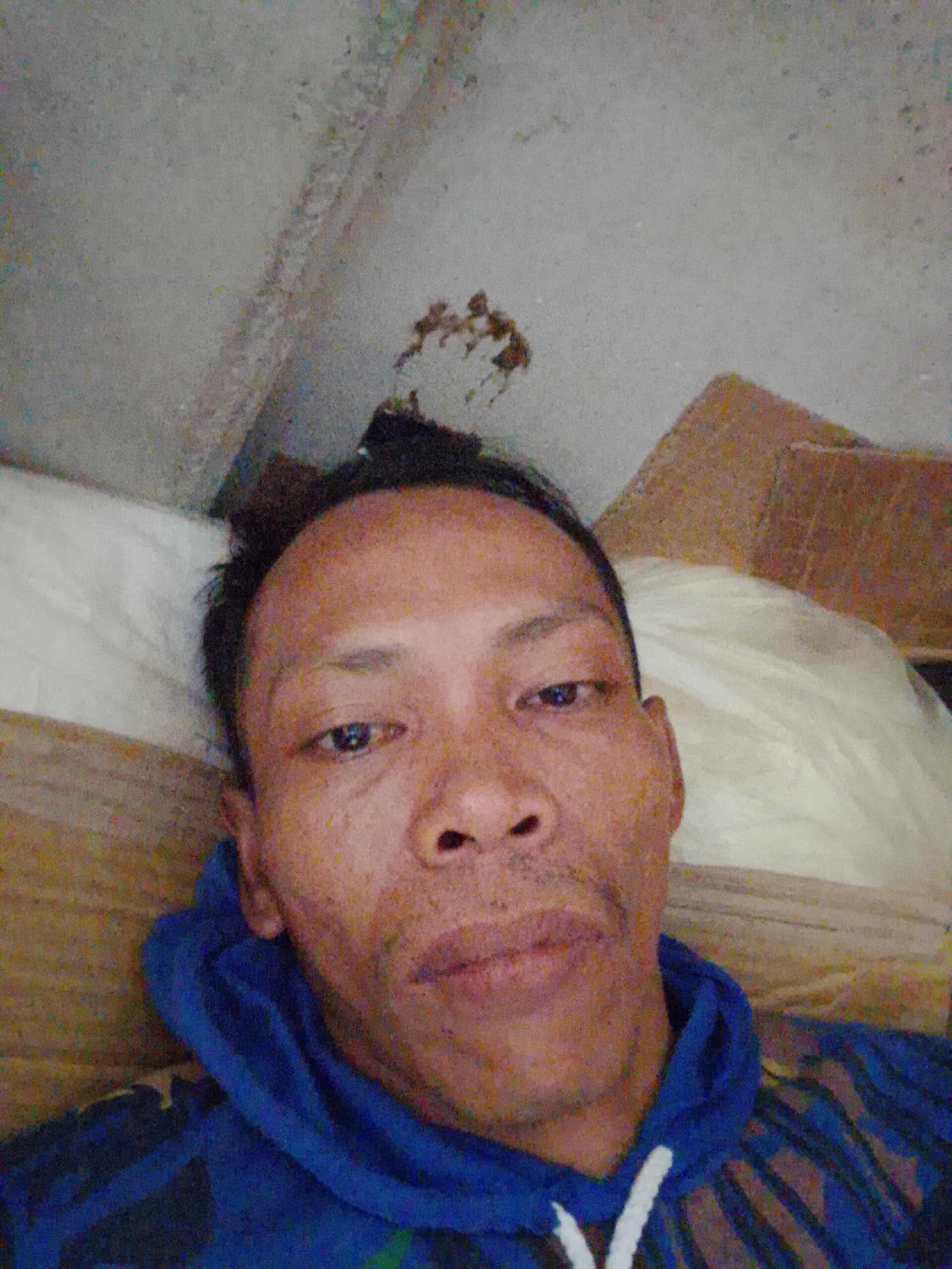 A man is lying down, facing the camera, in what appears to be an indoor space with unfinished or deteriorating walls. He is wearing a blue hoodie with a white V shape visible at the front. Behind him, there's a white pillow or cushion and what seems to be a cardboard box. Based on the prompt, this image originates from Bukit Ketok, Indonesia.
