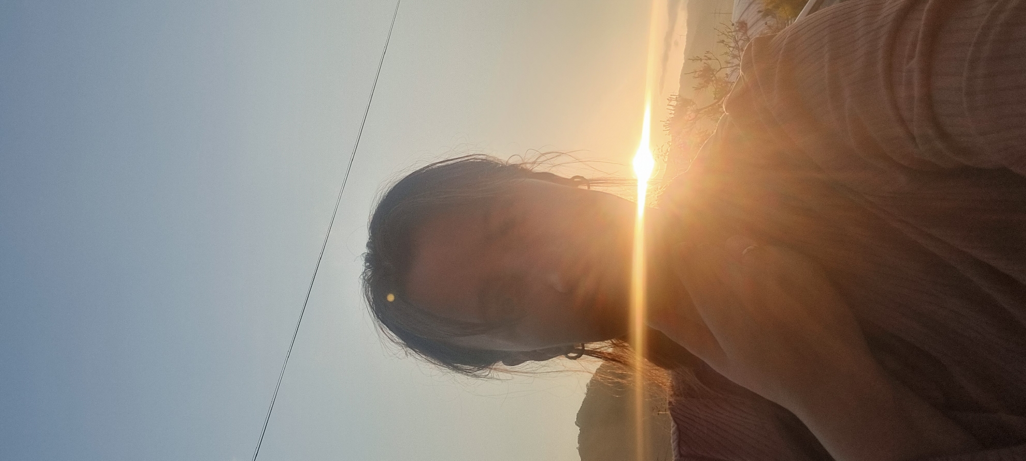 A person with dark hair faces the camera, their features obscured by intense sunlight. The background consists of a pale blue sky and landscape with a mountain silhouette in the distance. The setting sun casts a strong glare, making it difficult to discern details. A thin black cable stretches across the upper left portion of the image. The picture was taken in Dunnae-myeon, South Korea.