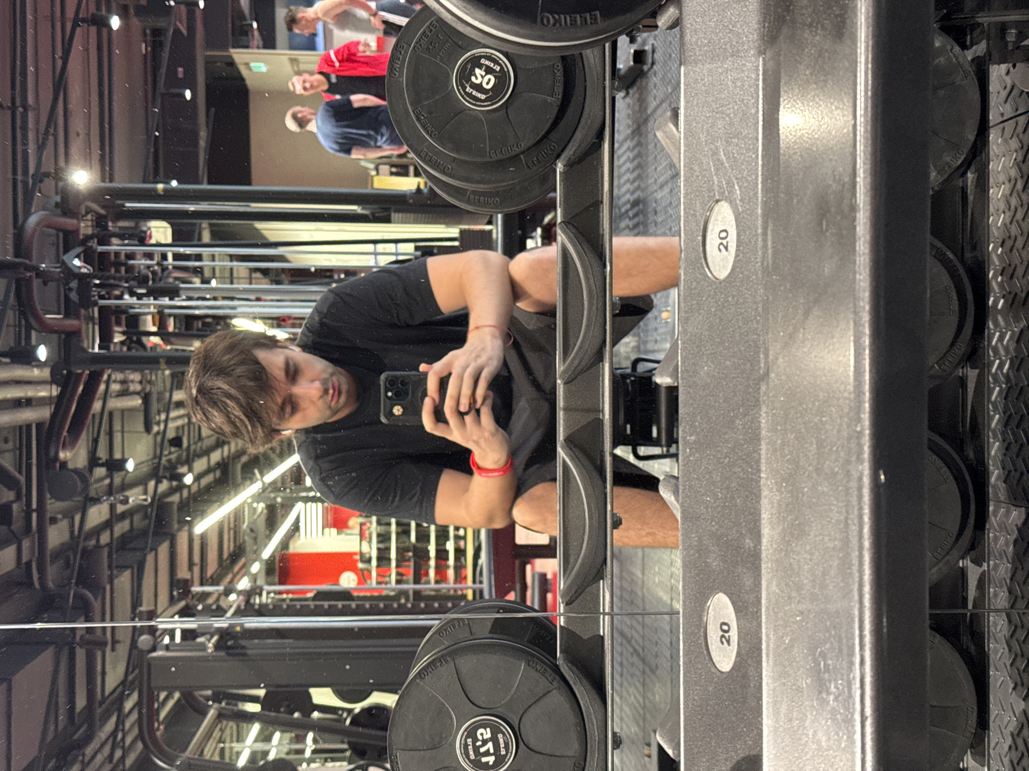 The image shows a mirror reflection of a man in a gym. He is wearing a black t-shirt and shorts. He is holding a smartphone in his right hand and appears to be taking a selfie. In the background, there are weight plates stacked on a rack. Some of the weight plates have "ELEIKO" and "20" written on them. The gym appears to be well-lit with fluorescent lights. The overall mood is casual and focused on fitness. The time of day is not specified, but the indoor lighting suggests it could be any time. The location cues point to a gym, possibly in Moscow, Russia, based on the provided context. The reflection shows other gym equipment and people in the background, suggesting a busy workout environment. The man's expression is neutral, focused on taking the photo.