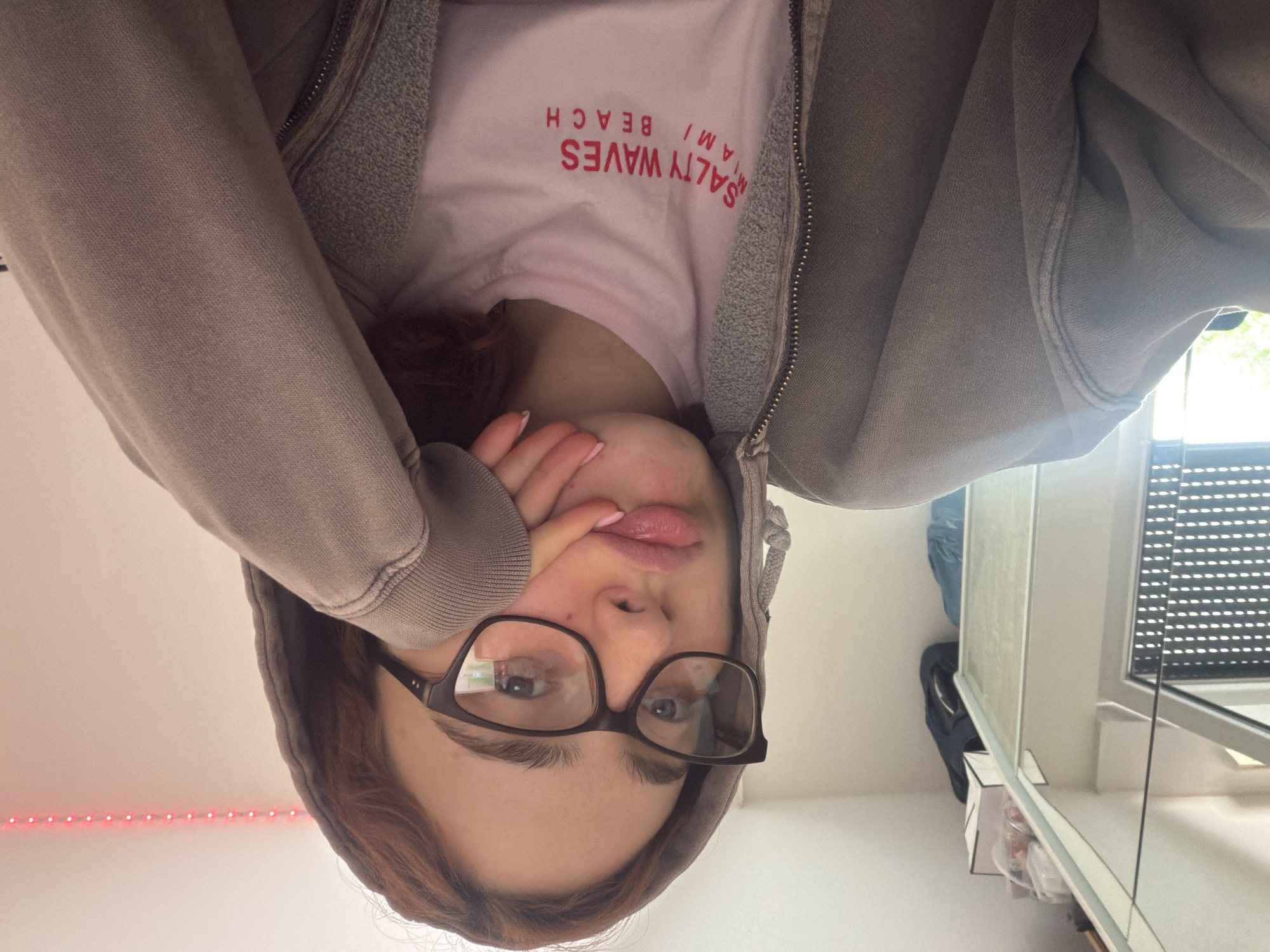 The image is a close-up, selfie-style photograph of a young woman wearing glasses and a grey hooded sweatshirt. She has her hand resting on her chin, and her lips are slightly pursed. A pink t-shirt with red text that reads "SALTY WAVES MIAMI BEACH" is visible underneath the sweatshirt. The background appears to be an indoor setting, possibly a room, with a white wall and a string of red LED lights. There is a window with blinds visible on the left side of the frame, and some shelves with objects are reflected in a mirrored surface. The lighting suggests it is daytime. This image is not associated with Strasbourg, France.