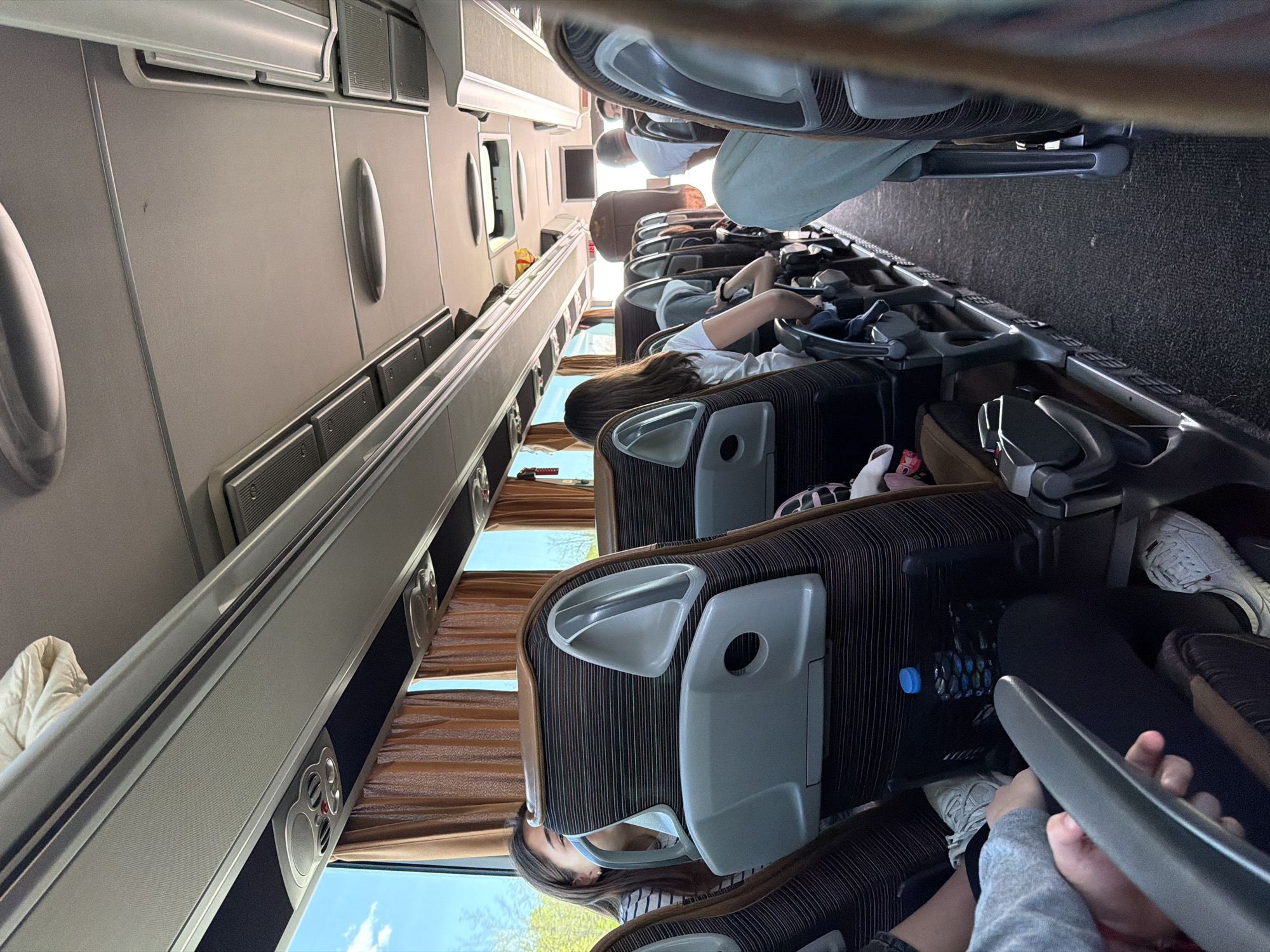 The image depicts the interior of a bus with passengers seated. Passengers are in the foreground and middle ground. The interior is dimly lit, with overhead compartments running along the left side of the frame. Brown curtains cover the windows. Seats are brown with a striped pattern and grey headrests. Passengers are in varying states of relaxation, with some looking out the windows. A water bottle is visible next to one of the seats. The top portion of the image shows luggage racks. The location in the city of Yazdach, Bulgaria, is not discernible from the image.