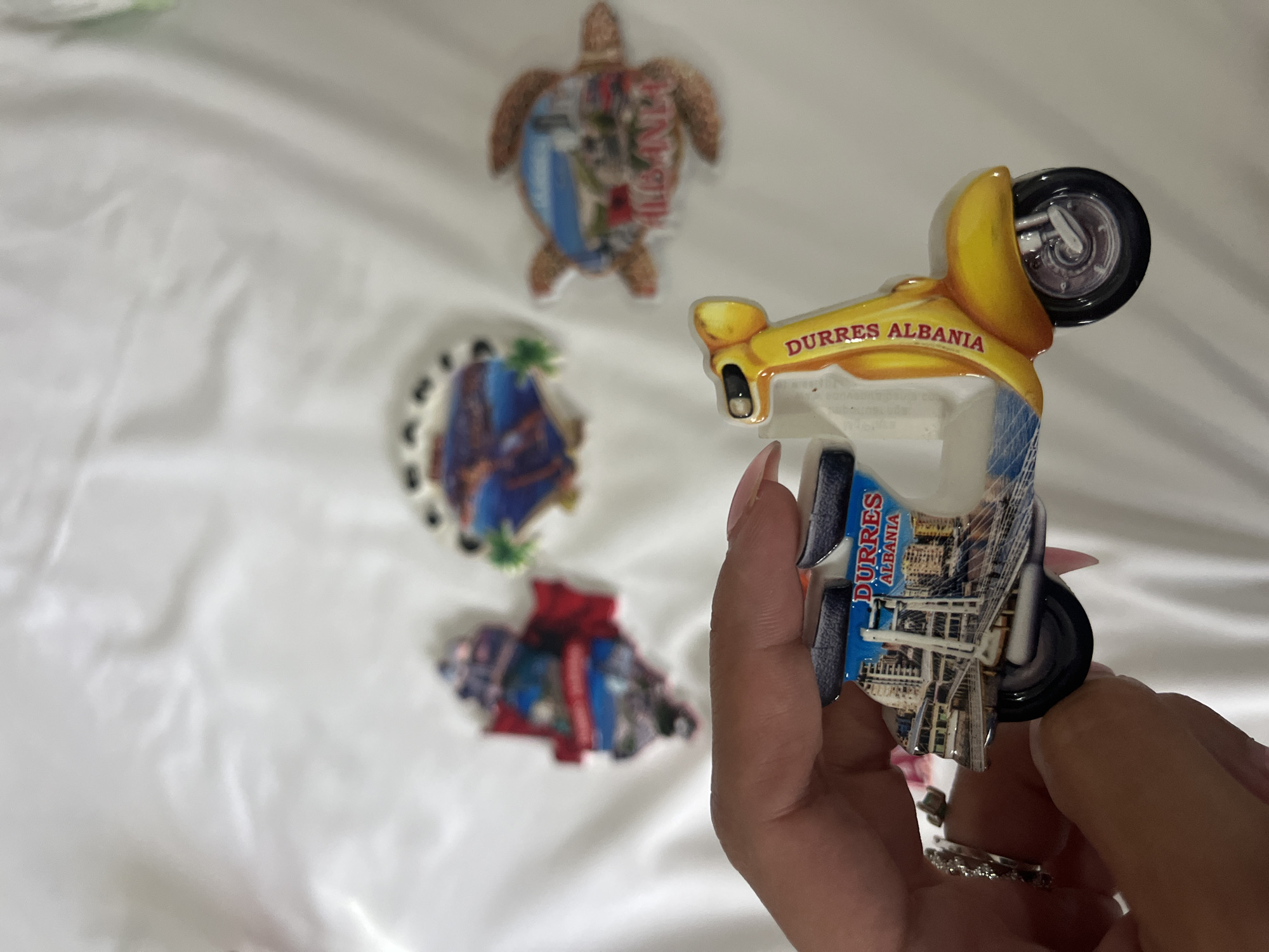 A person's hand with painted nails and rings is holding a yellow scooter-shaped magnet. The magnet depicts the city of Durrës, Albania, featuring a bridge and buildings. In the background, on a white, crinkled surface, are three other magnets: one shaped like Albania with a city scene and red flag, another resembling a round plate with a cityscape and palm trees, and a third shaped like a turtle with a cityscape and the word "ALBANIA" printed on it.