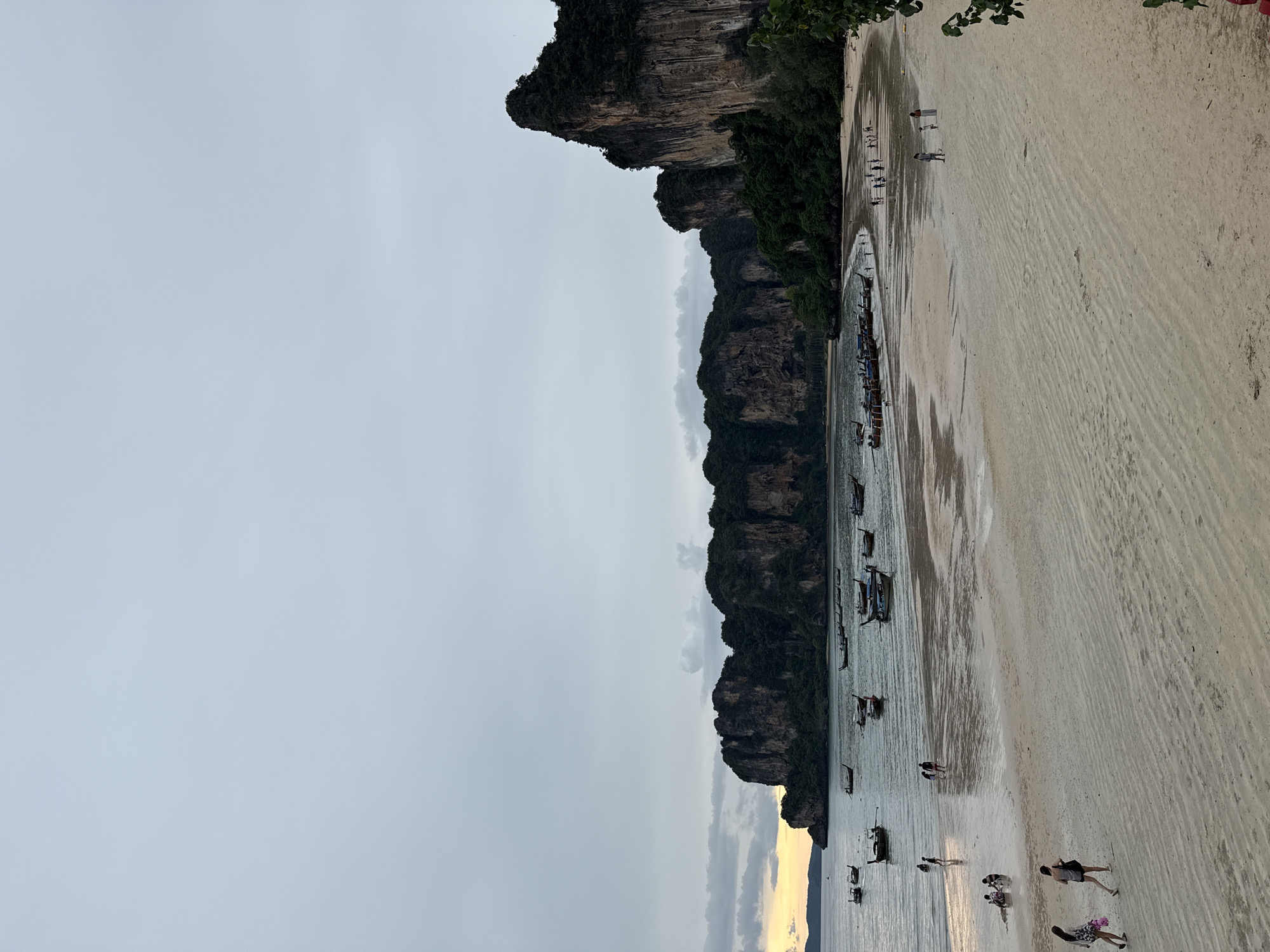 The image depicts a beach scene in Ban Ao Nang, Thailand, during what appears to be dusk or overcast conditions. Towering, verdant limestone cliffs dominate the background, with numerous long-tail boats moored in the calm, shallow water. The sandy beach, marked by textured ripples from the receding tide, is sparsely populated by individuals walking or sitting. A few people are visible near the water's edge, and others are scattered further up the beach. The sky is a pale, overcast gray, with a faint hint of warm light on the horizon to the far left, suggesting the setting or rising sun. The overall atmosphere is serene and quiet.