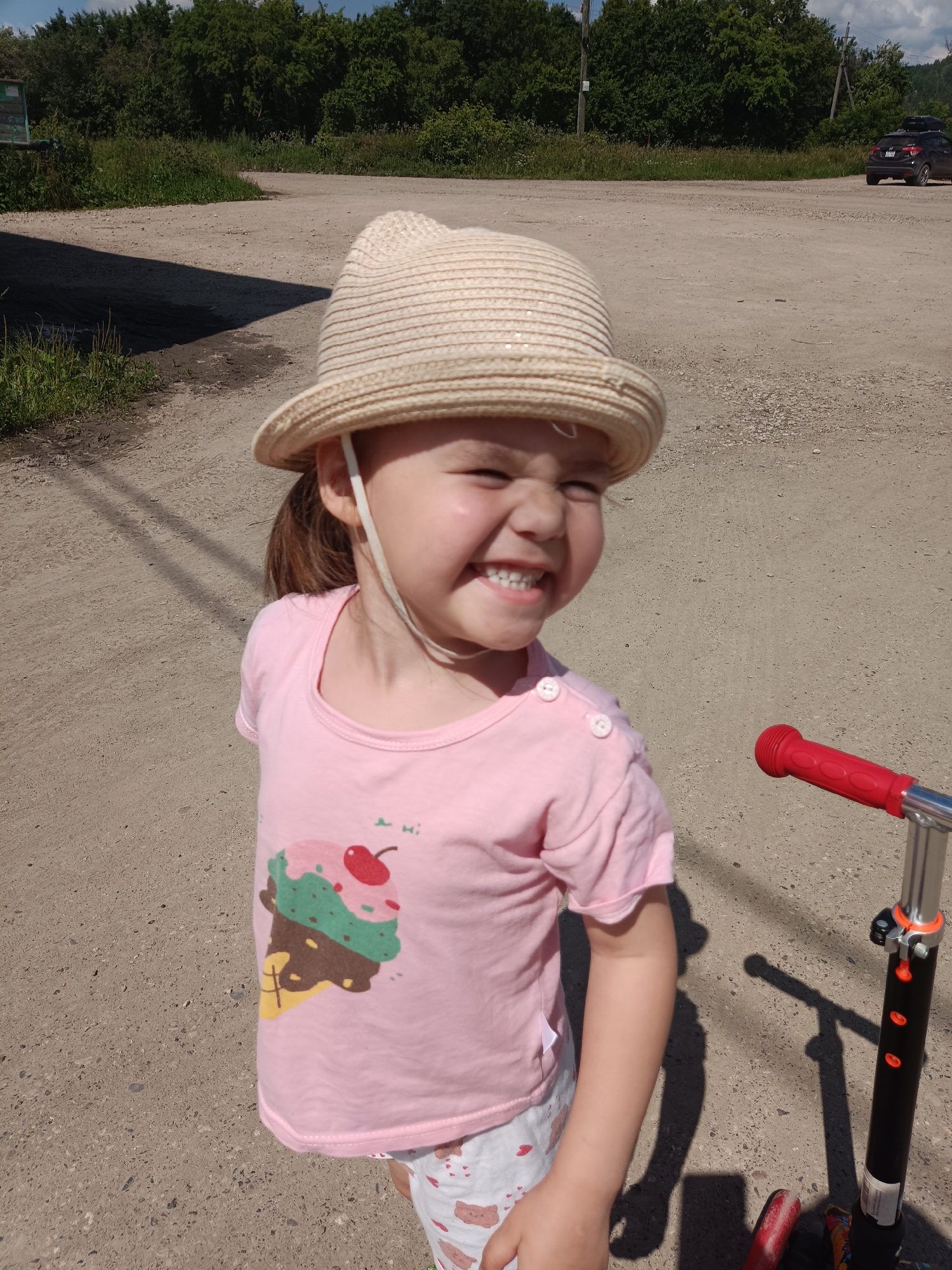 In the city of Posad, Russia, a young girl with brown hair, wearing a straw hat and a pink t-shirt with an ice cream cone graphic, smiles broadly. She is standing on a dirt road next to a black and red scooter. Trees and a dark-colored car are visible in the background. The scene appears to be outdoors during the daytime.