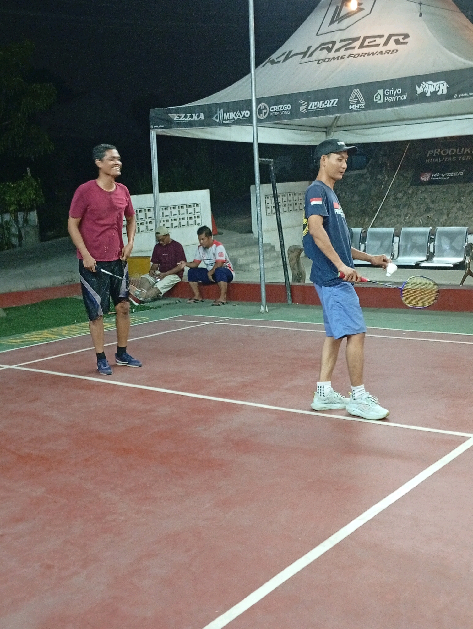 A group of men are gathered on a badminton court in what appears to be an outdoor recreational facility at night. Two men are actively playing badminton. One man, wearing a maroon t-shirt and dark shorts, stands on the court holding a badminton racket in his left hand. He is looking to his right with a smile. Another man, wearing a dark blue t-shirt with an Indonesian flag emblem and light blue shorts, stands on the court holding a badminton racket and a shuttlecock. He is looking down and to his right. In the background, two men are seated on a low wall. One man wears a maroon t-shirt and light shorts, while the other wears a white shirt with red accents and dark shorts. They appear to be watching the game. Behind them, there is a white tent with logos and text, including "KHAZER COME FORWARD". Further in the background are rows of empty seating. The court surface is red with white lines, and there is a green area with yellow markings to the left. The lighting suggests it is nighttime, and the overall environment seems to be a sports complex. The image was taken in Caringin, Indonesia.
