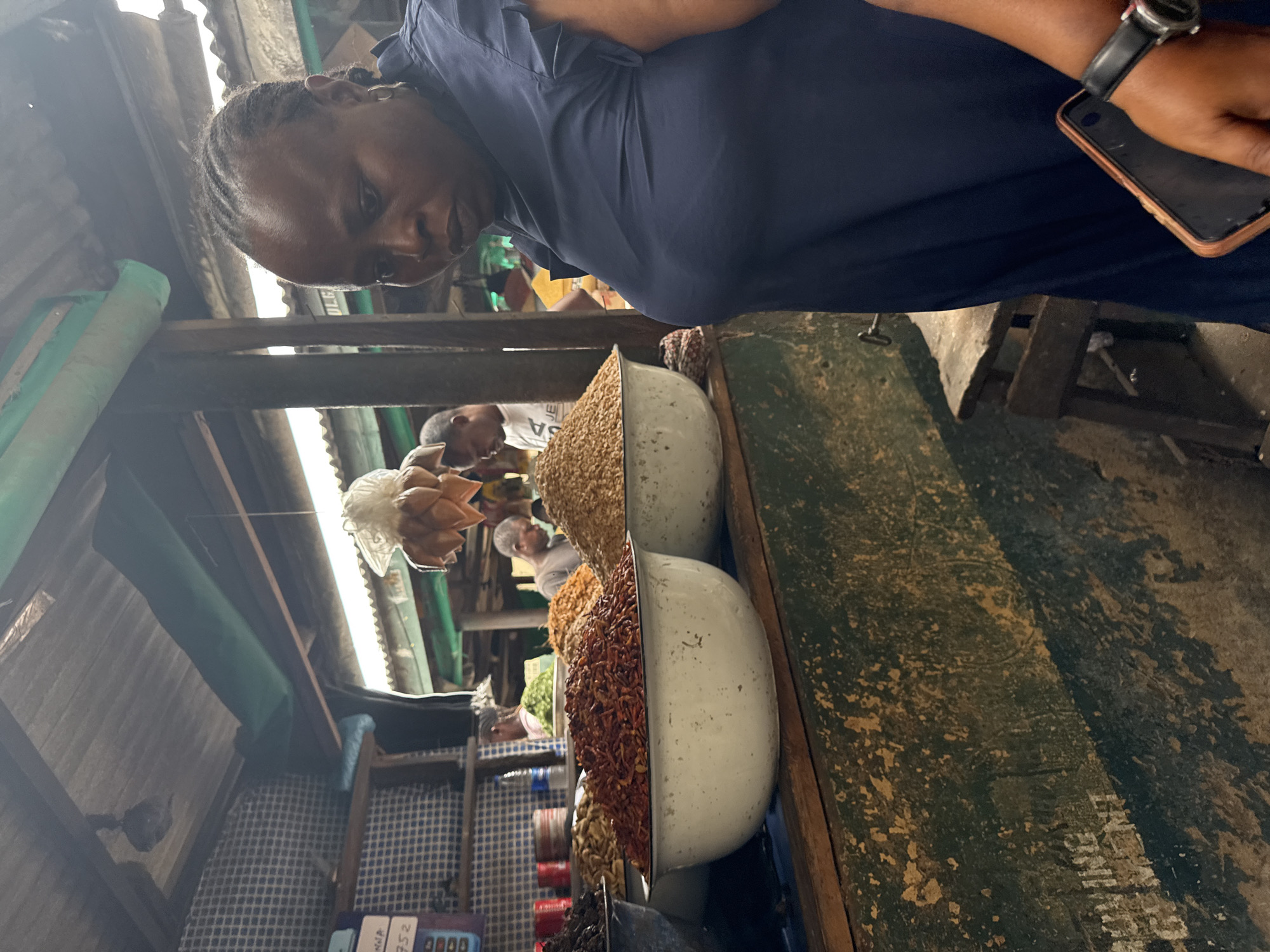 A dark-skinned woman with braided hair and a navy blue dress stands in a market. She is holding a phone in her right hand. In front of her, there are several bowls of dried goods, including what appears to be dried shrimp or chili flakes in the foreground. In the background, other people are visible, and the market appears to be covered by a corrugated metal roof. The scene is likely in Lagos, Nigeria, given the presence of a market and the overall visual context.